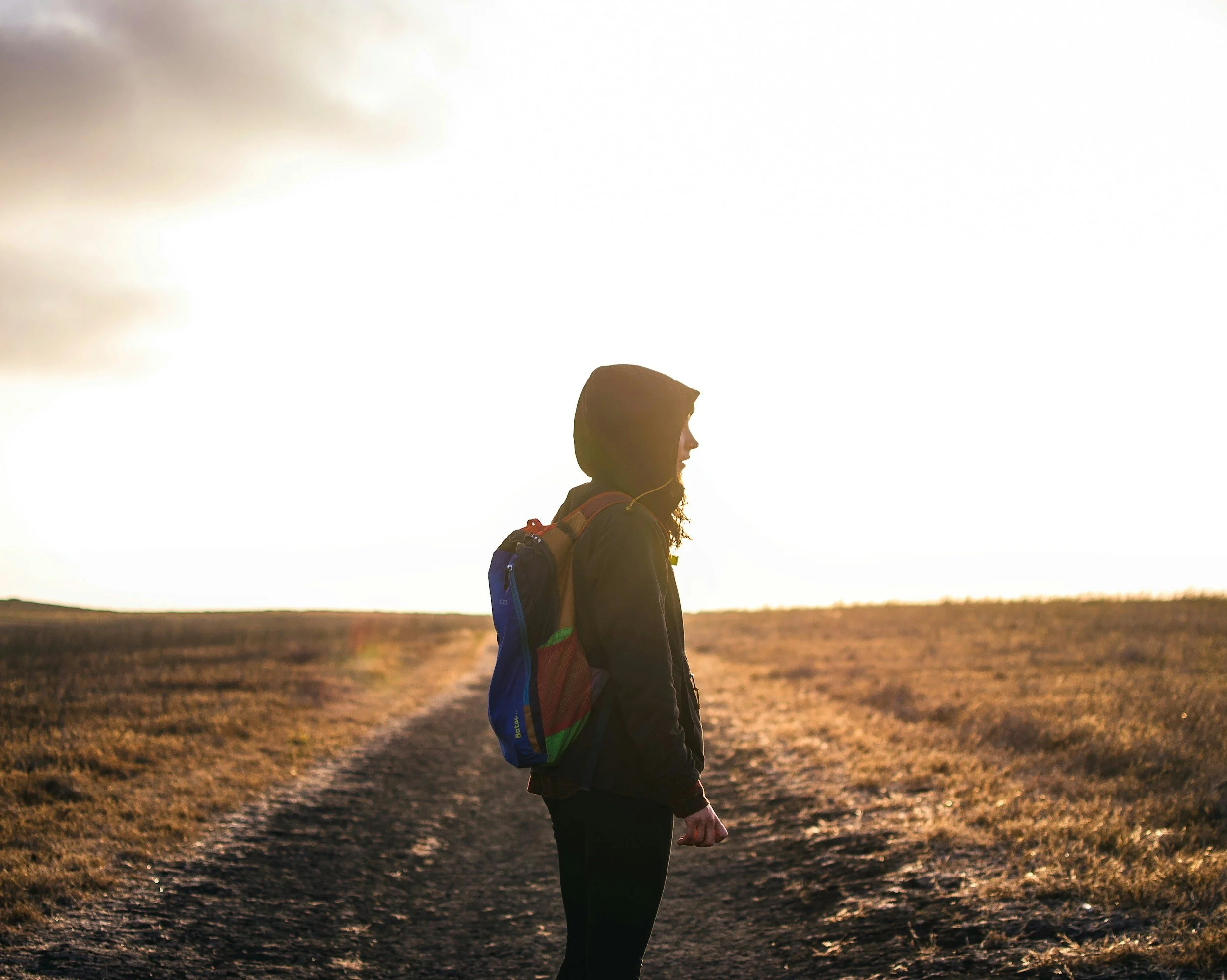 A woman in a black jacket and colorful backpack, wearing a hood, standing on a dirt path in a field during sunset, facing to the right.