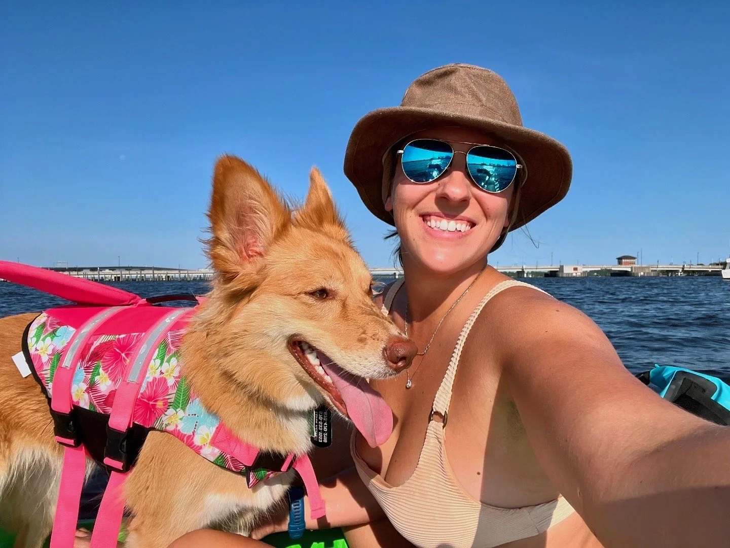 A woman smiling for a selfie with her dog on a boat, wearing sunglasses and a wide-brimmed hat, with water and a bridge in the background.