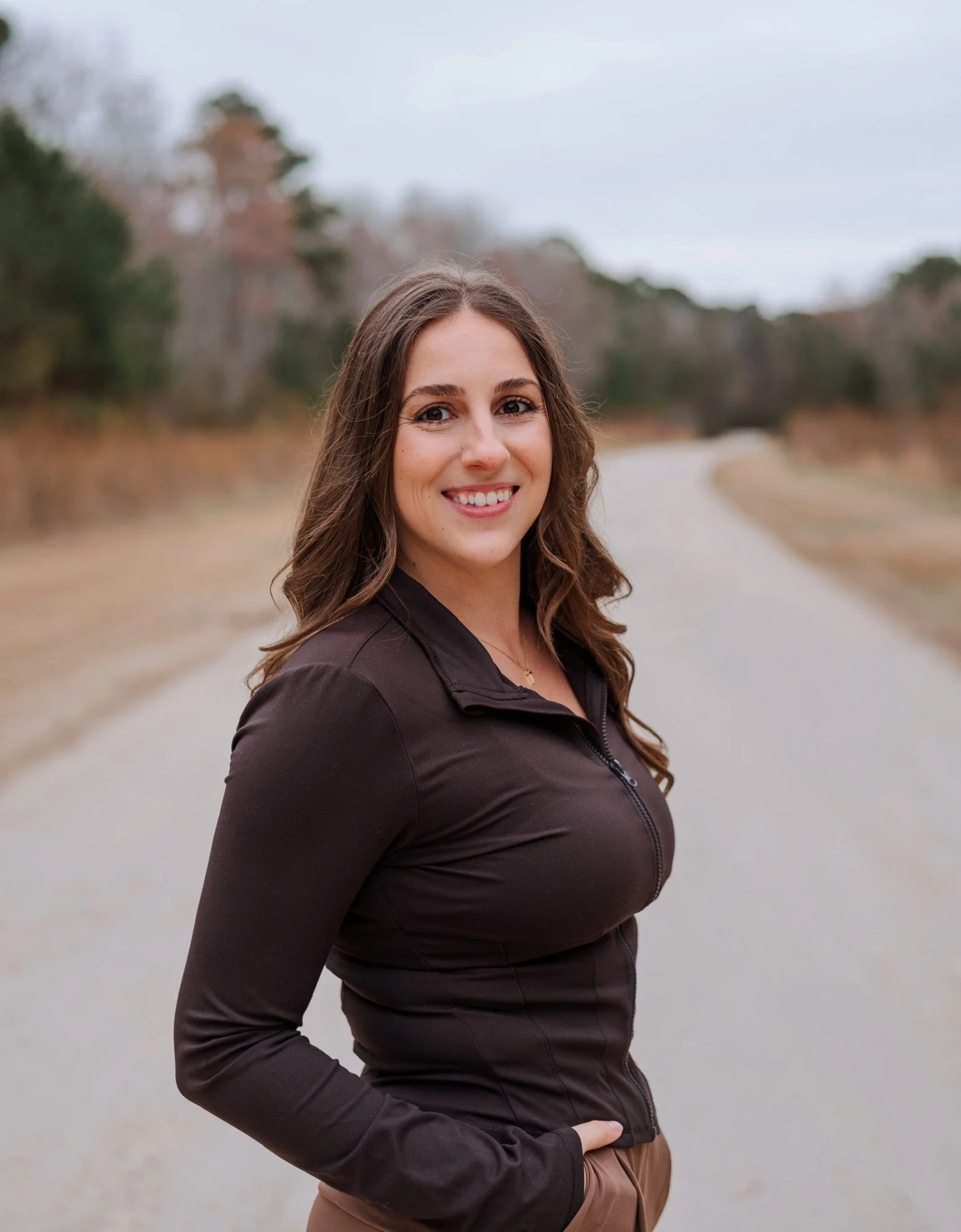 A woman with long brown hair, smiling, wearing a black zip-up jacket and brown pants, standing outdoors on a cloudy day near a curving road with trees in the background.