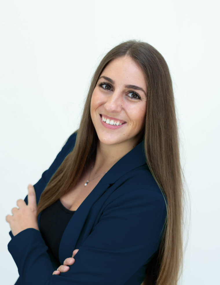 A young woman with long brown hair smiling, wearing a dark blazer and a black top, standing against a plain white background.