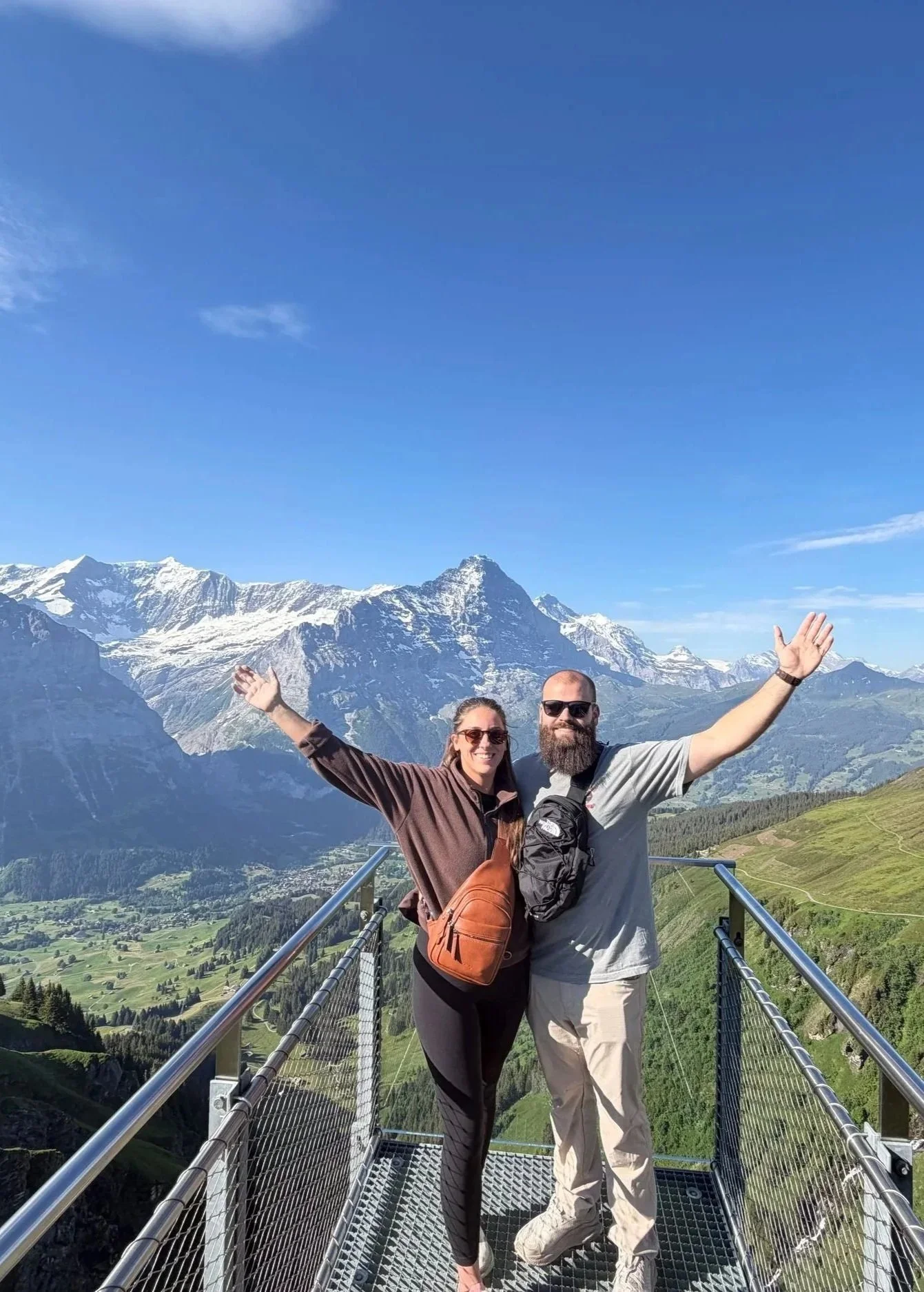 A man and woman standing on a metal suspension bridge in front of snow-capped mountains, both with arms raised. The woman is smiling, wearing sunglasses, a brown jacket, black pants, and a orange backpack. The man is also smiling, wearing sunglasses, a gray t-shirt, tan pants, and has a beard. The landscape behind them shows lush green valleys, fields, and a clear blue sky.