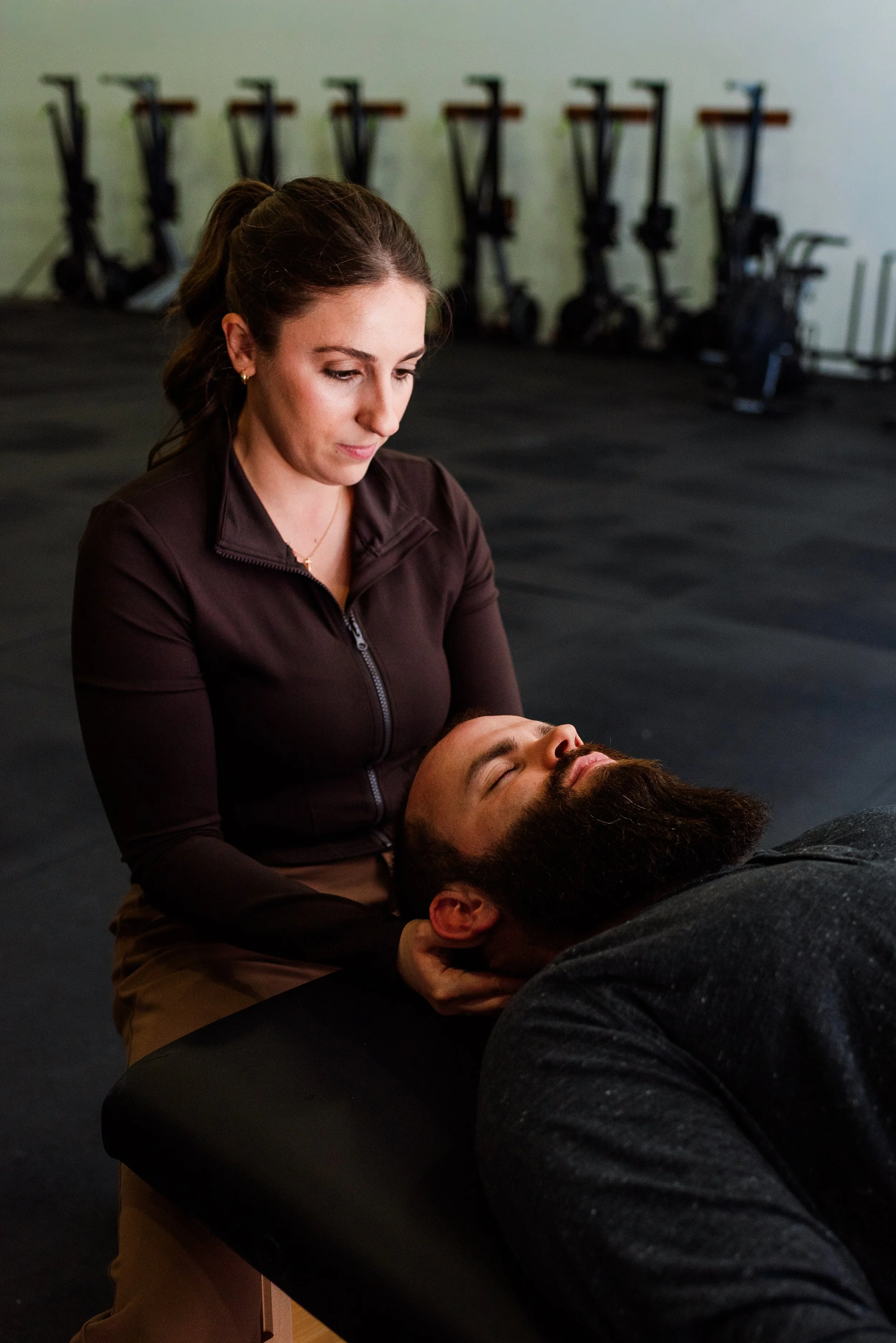 A woman performing a chiropractic adjustment or therapy on a man lying on a treatment table in a gym or therapy room, with exercise bikes in the background.