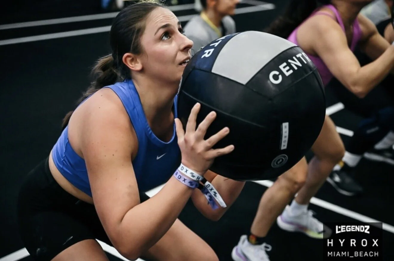 A woman in a blue athletic top holding a medicine ball while performing a workout in a gym with other women.