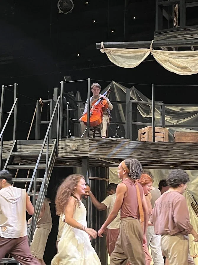 Scene from a theater performance featuring a musical setting. A person is seated on a balcony playing a bright orange cello, while a group of performers are dancing and interacting below on stage. The stage background has draped fabric and set pieces, creating an artistic and theatrical atmosphere.