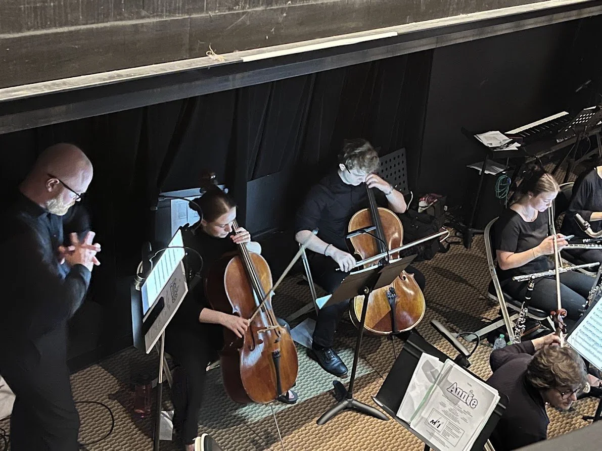 Musicians in black clothing practicing in a rehearsal space, playing a cello, flute, and other instruments, with sheet music on stands.