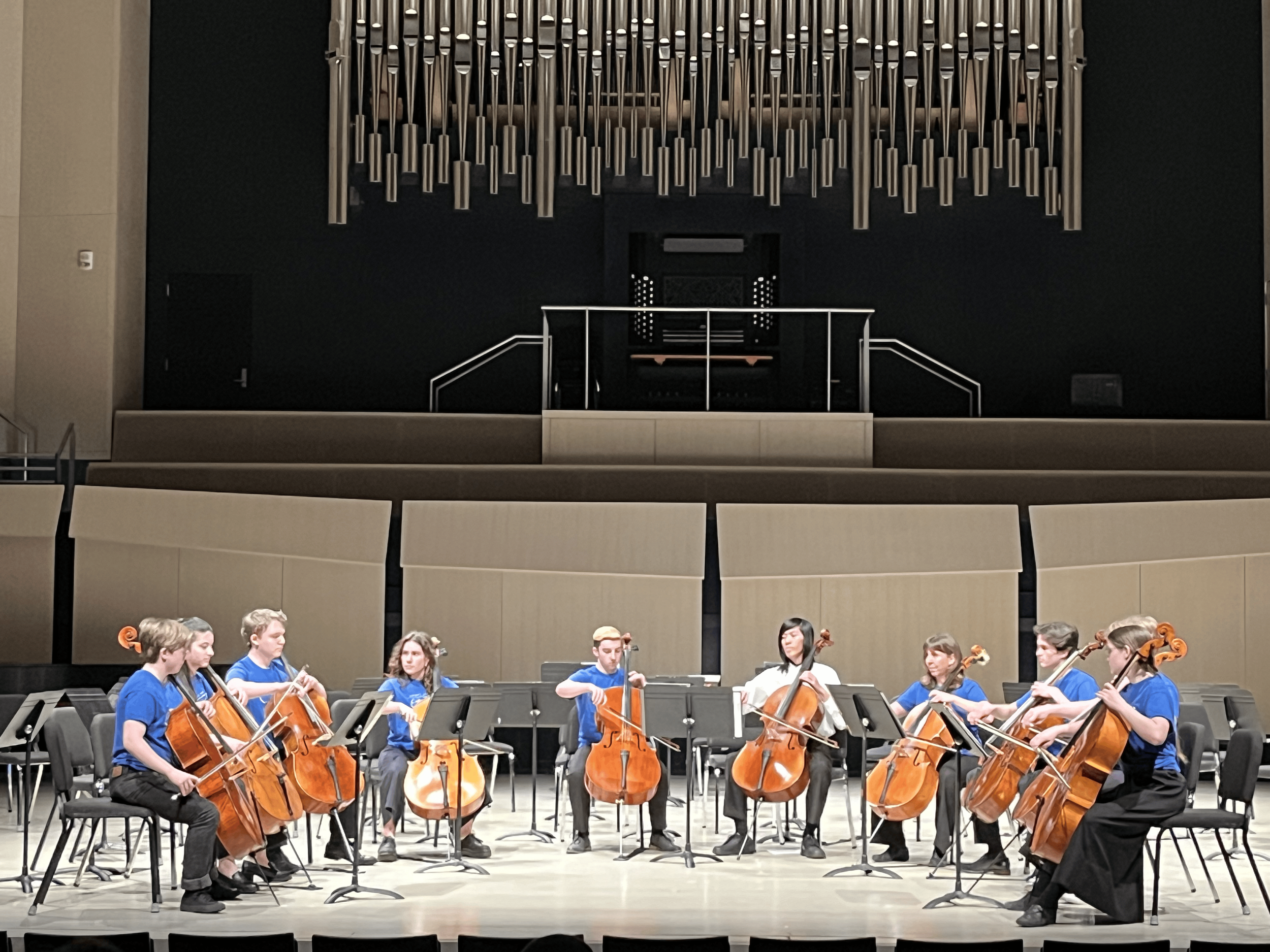 A group of musicians playing string instruments, such as cellos and violins, on stage during a performance, with a large pipe organ in the background.