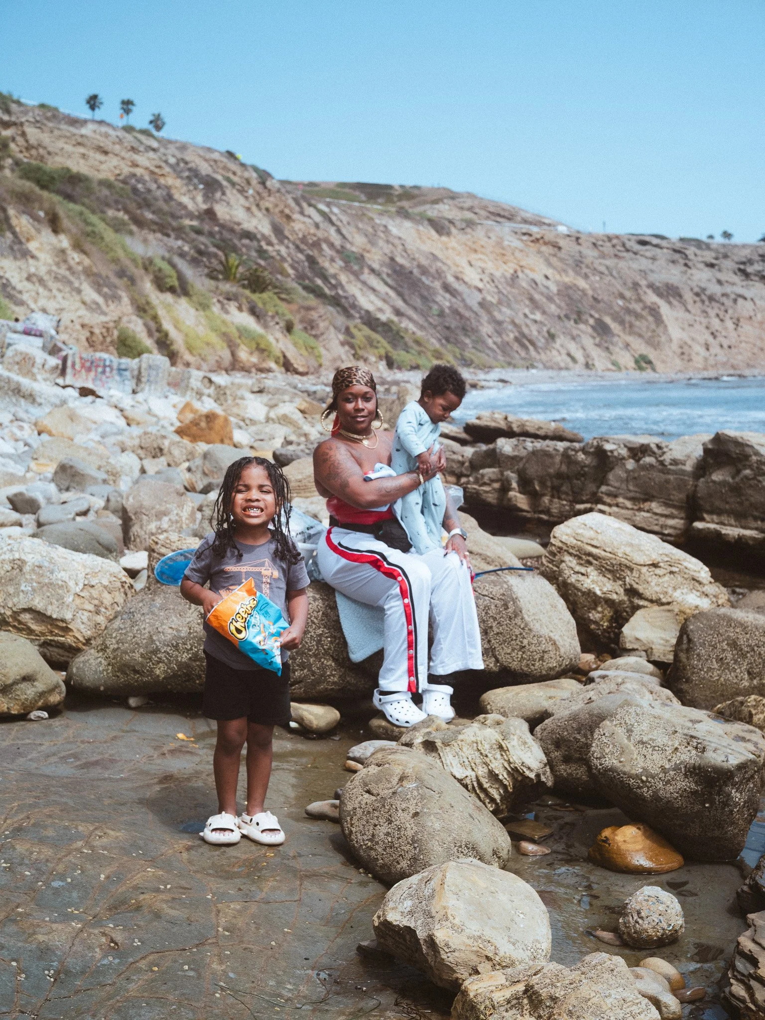 A family of three by the rocky shoreline of a beach, with hills and sparse vegetation in the background. The girl is holding a bag of Cheetos, smiling at the camera, wearing white sandals, black t-shirt, and shorts. The woman is sitting on a rock, holding a child, dressed in a red top and white pants with red and black stripes, and the man is standing behind them on the rocks, looking down, with dark curly hair, wearing a light blue shirt and patterned pants.