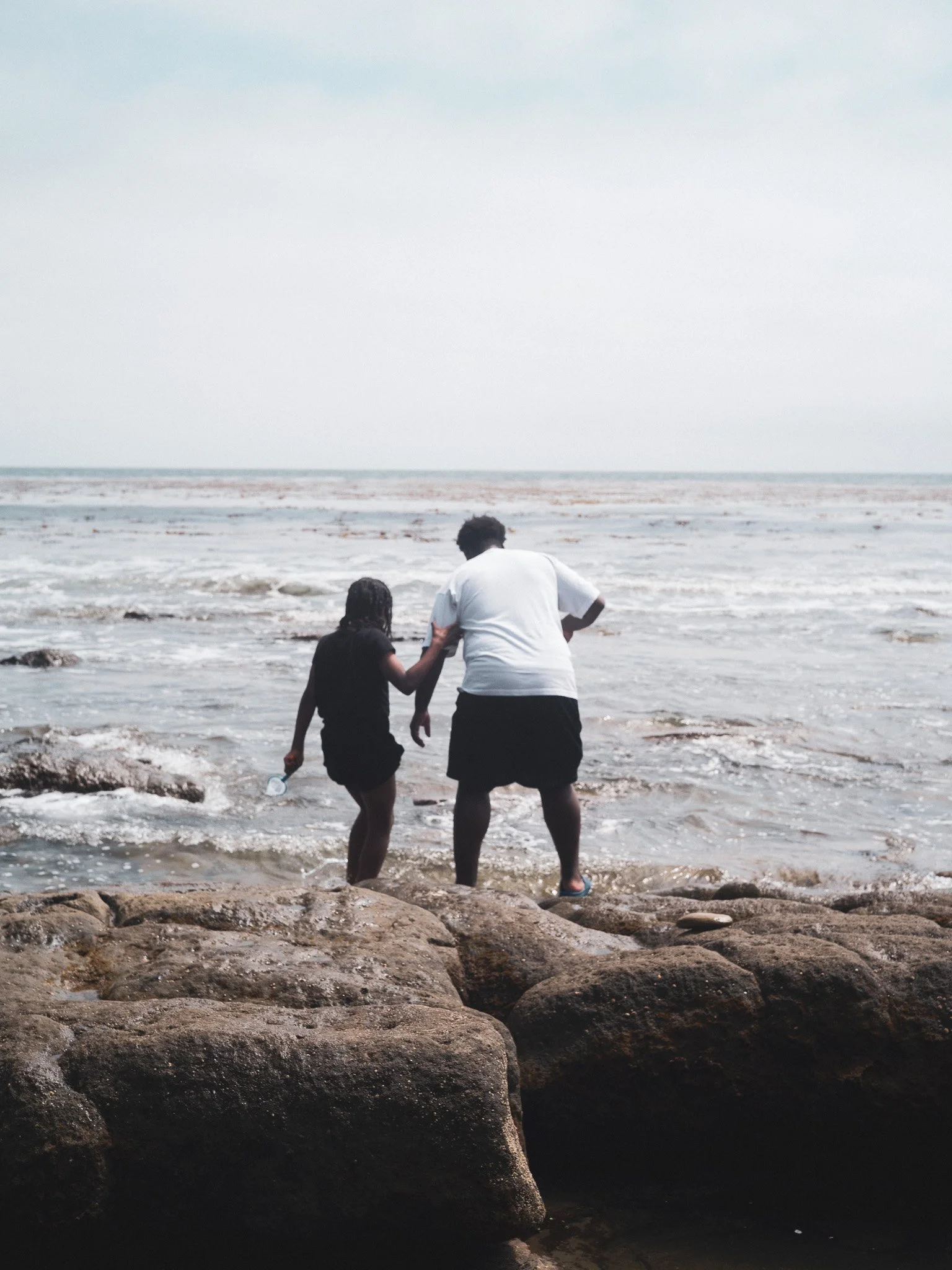 Two children standing on rocks at the edge of a beach with the ocean in the background.