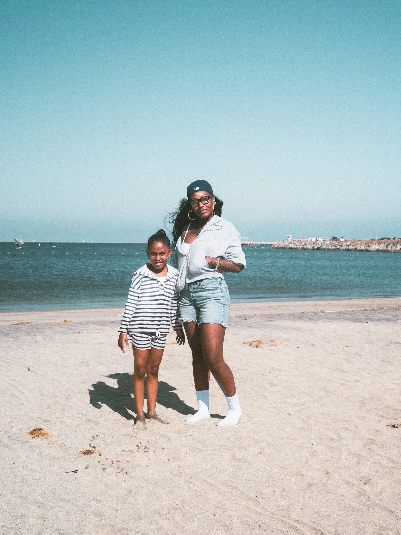 A woman and a young girl standing on a sandy beach near the water, with sailboats and a pier in the background. The woman is wearing a hoodie, shorts, white socks, and glasses, while the girl is dressed in a striped hoodie and shorts.