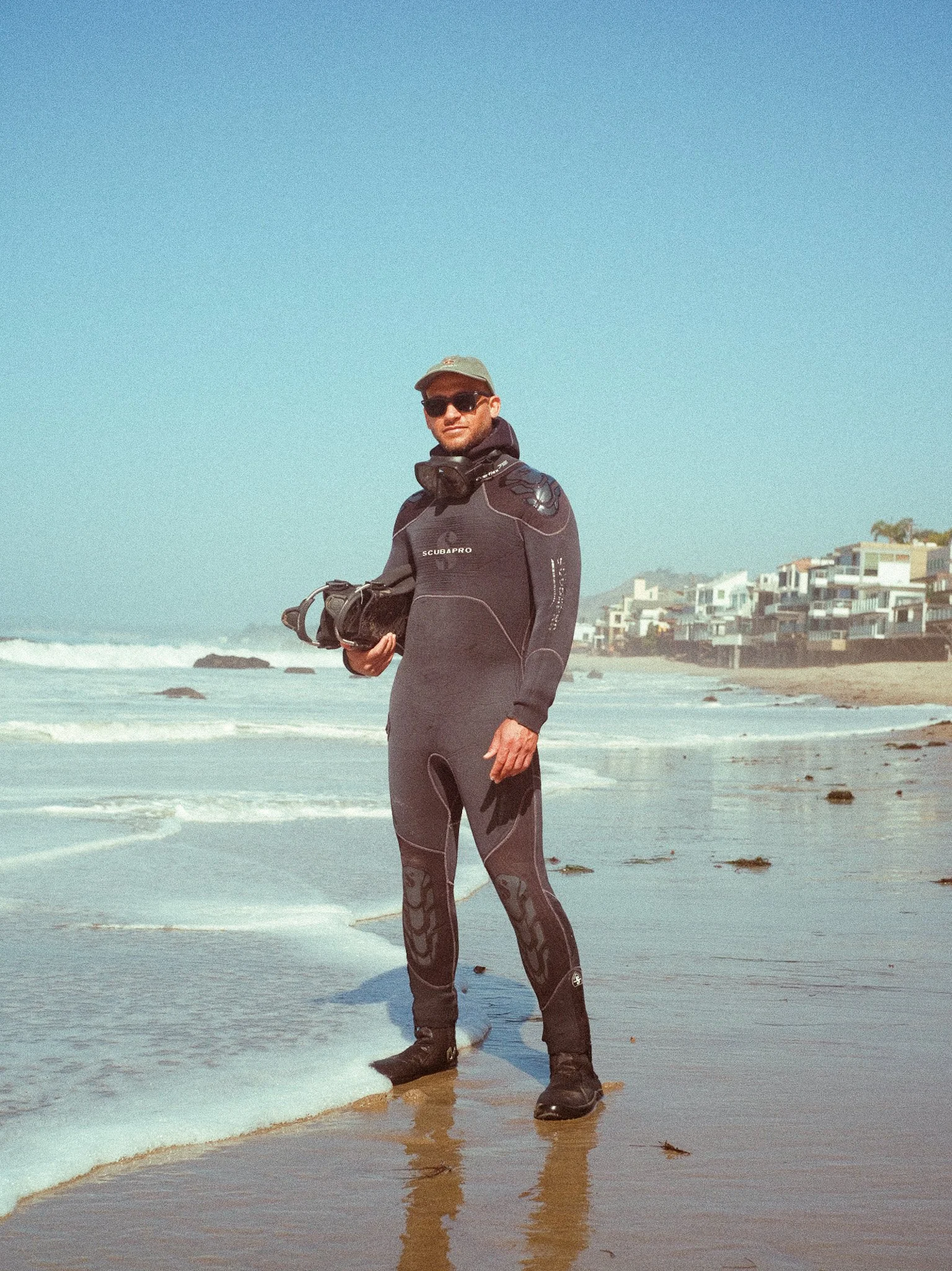 A man in a wetsuit and sunglasses standing in shallow water on a beach, holding a snorkel mask, with a row of modern beach houses in the background.