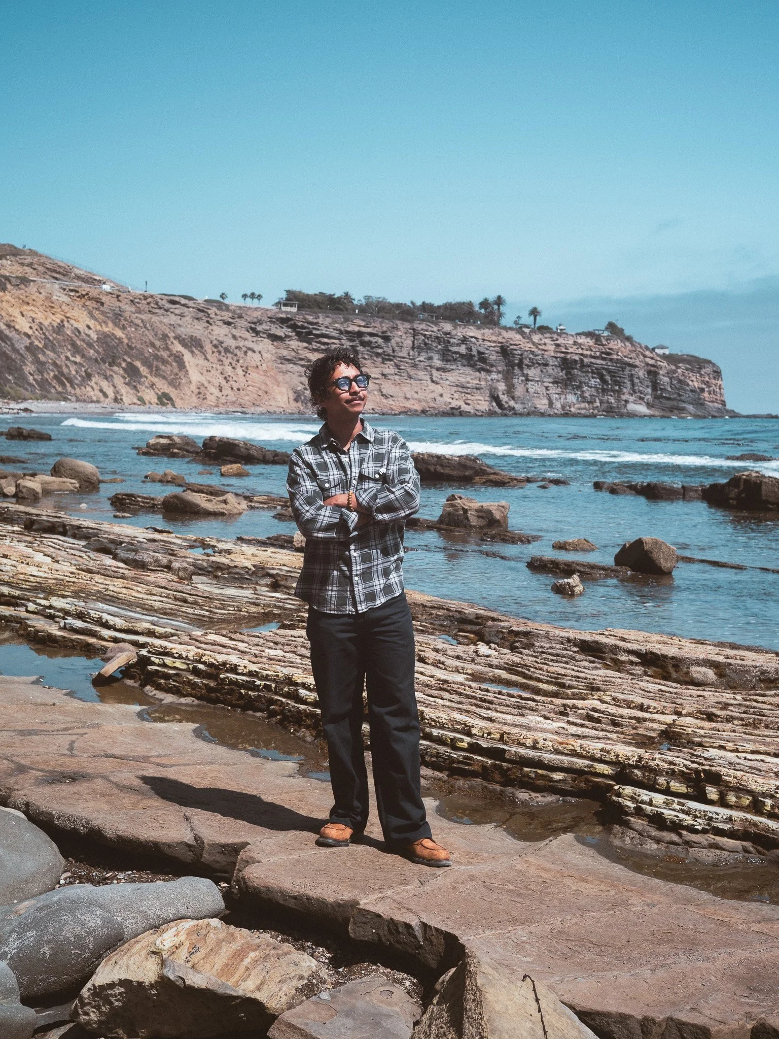 A person stands on a rocky shoreline with their arms crossed, wearing glasses, a plaid shirt, dark pants, and brown shoes. In the background, there are large rocks, ocean waves, cliffs, and a clear blue sky with some clouds.