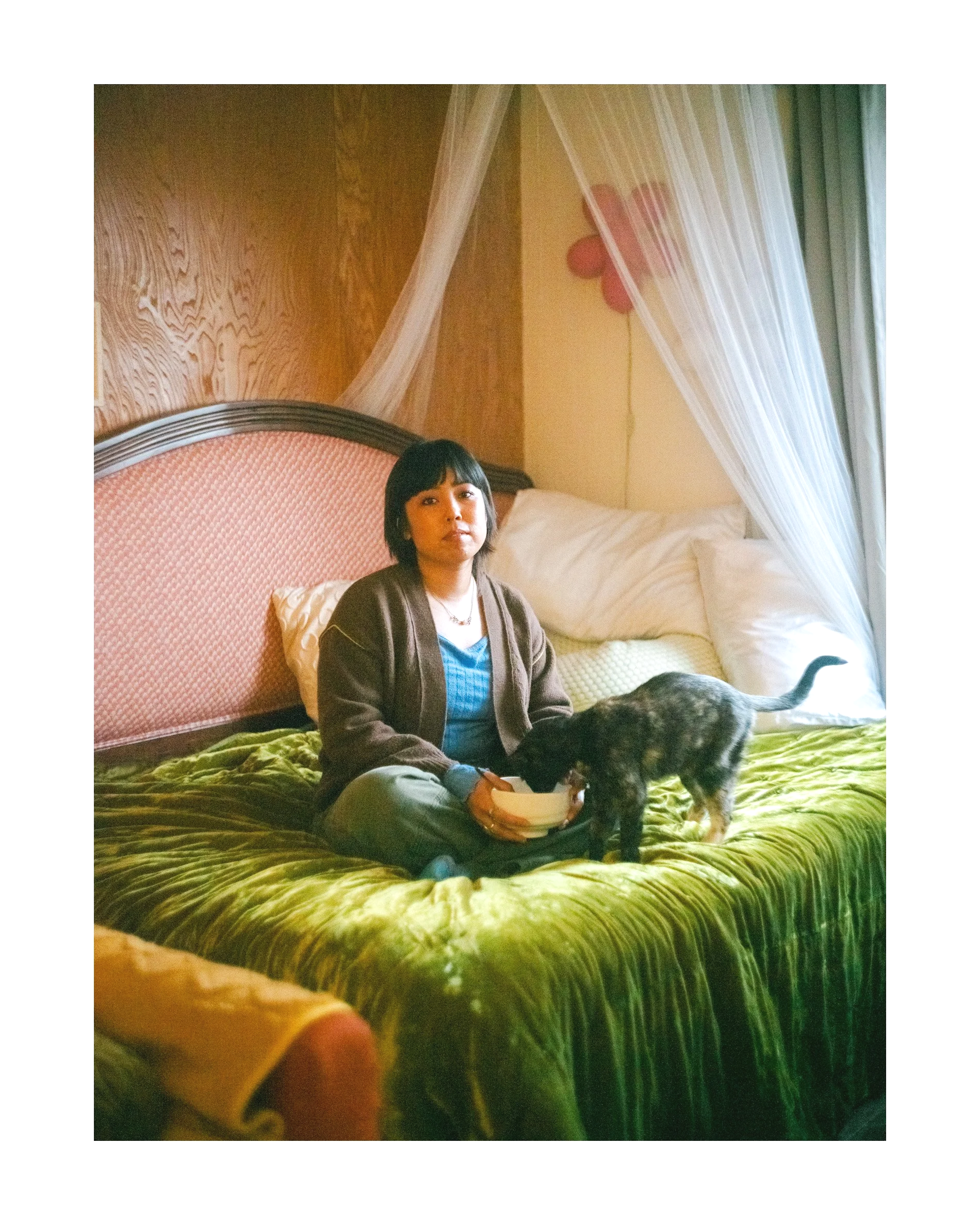 A woman sitting on a bed with green bedding, holding a bowl, with a dog nearby. The bed has a pink upholstered headboard, and sheer curtains draped above it. There is a pink flower decoration on the wall behind, and the room has wooden paneling and a