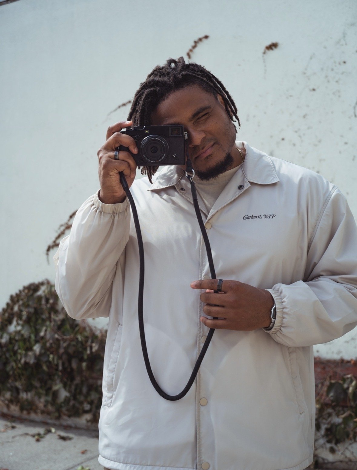 Man with dreadlocks taking a selfie using a camera, standing outdoors near a white wall with some dried plants and bushes in the background.