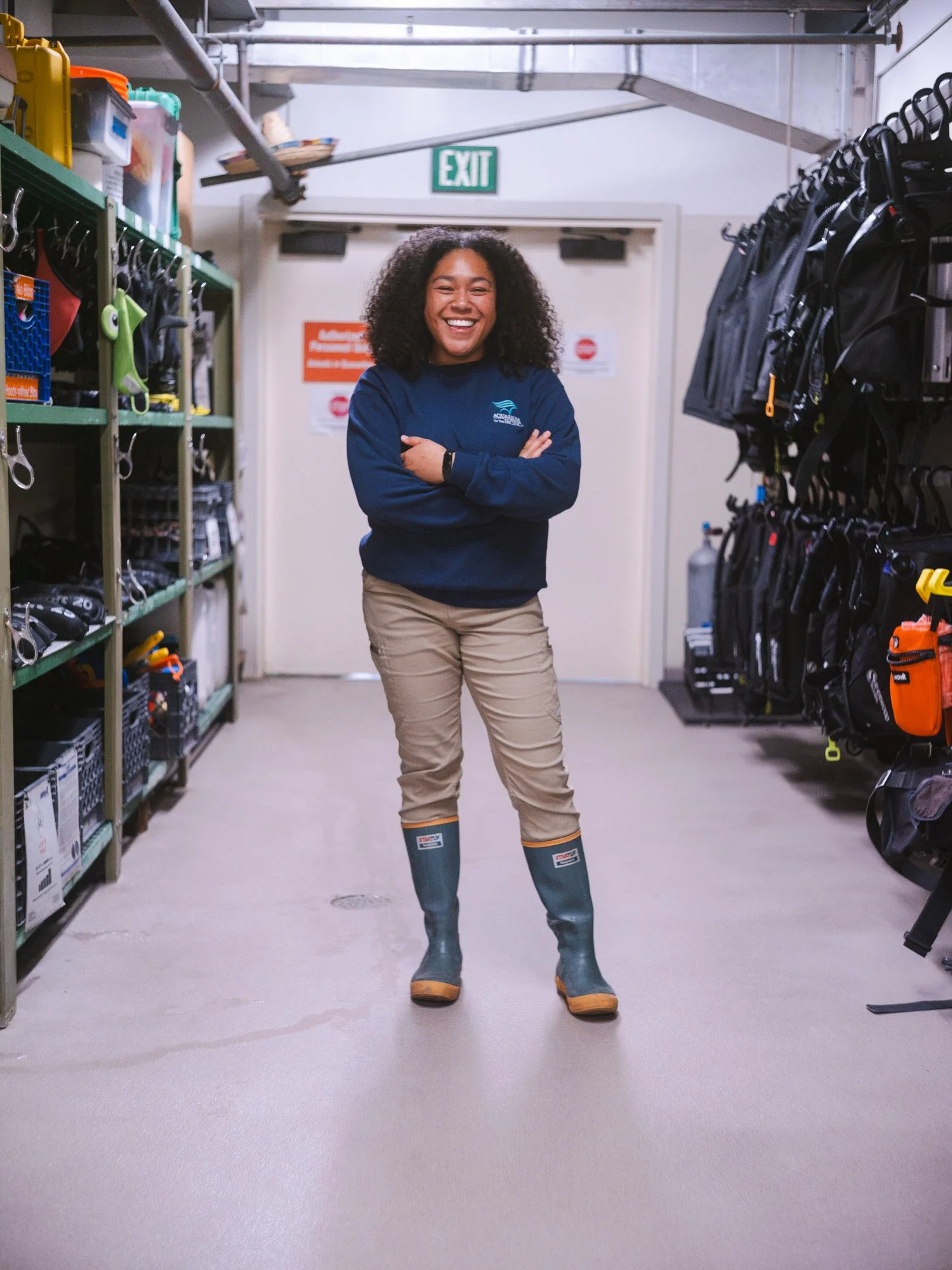 A woman standing in a storage room or workshop, smiling, with arms crossed, wearing beige pants, a navy blue long sleeve shirt, and blue rubber boots. Shelves with tools and equipment are on either side of her, and there is an exit sign above a door behind her.