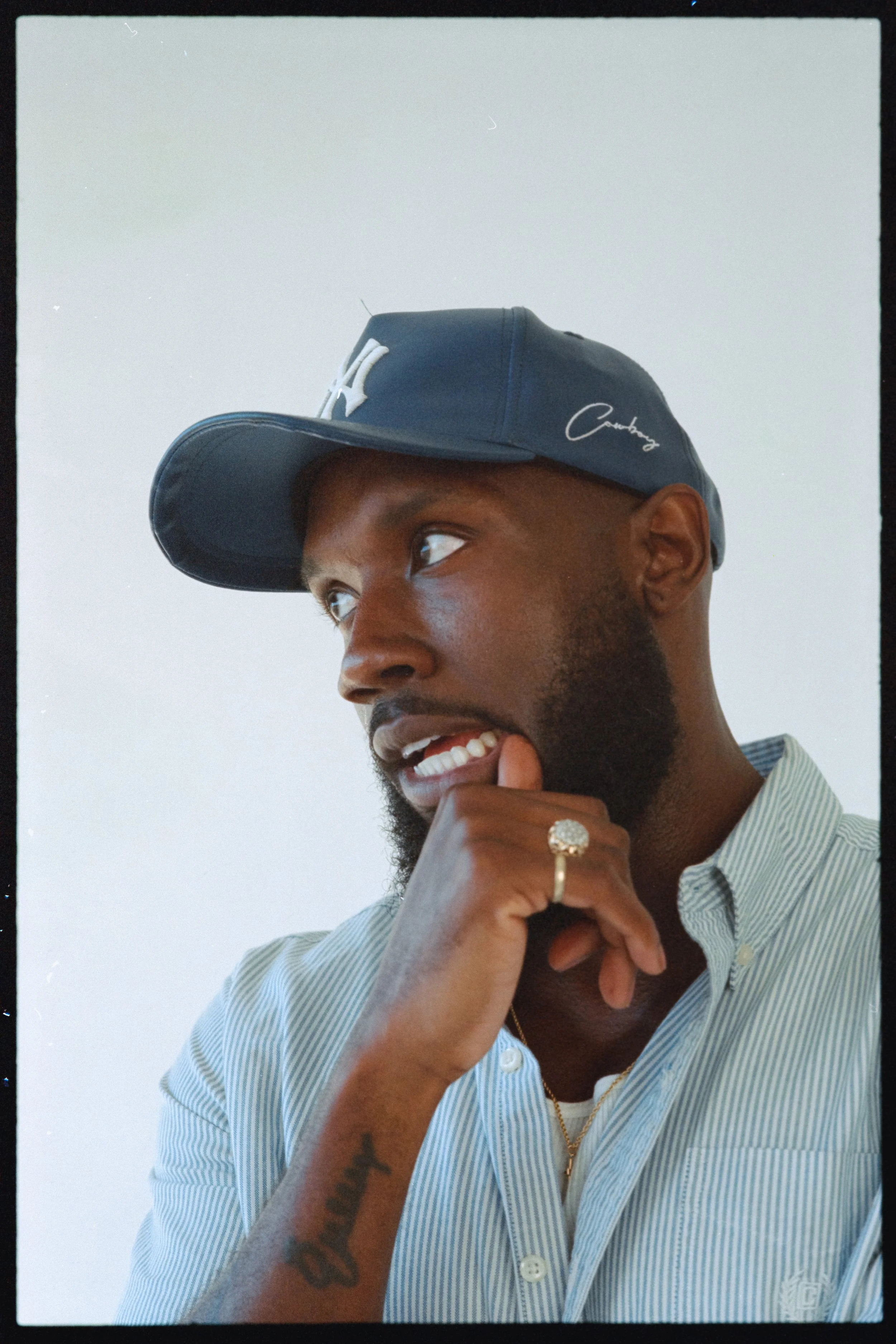 A young man with a short beard and dark skin, wearing a blue baseball cap with a white logo and signature, a light blue and white striped shirt, a gold chain, and a ring, is smiling with his tongue out and resting his chin on his hand.