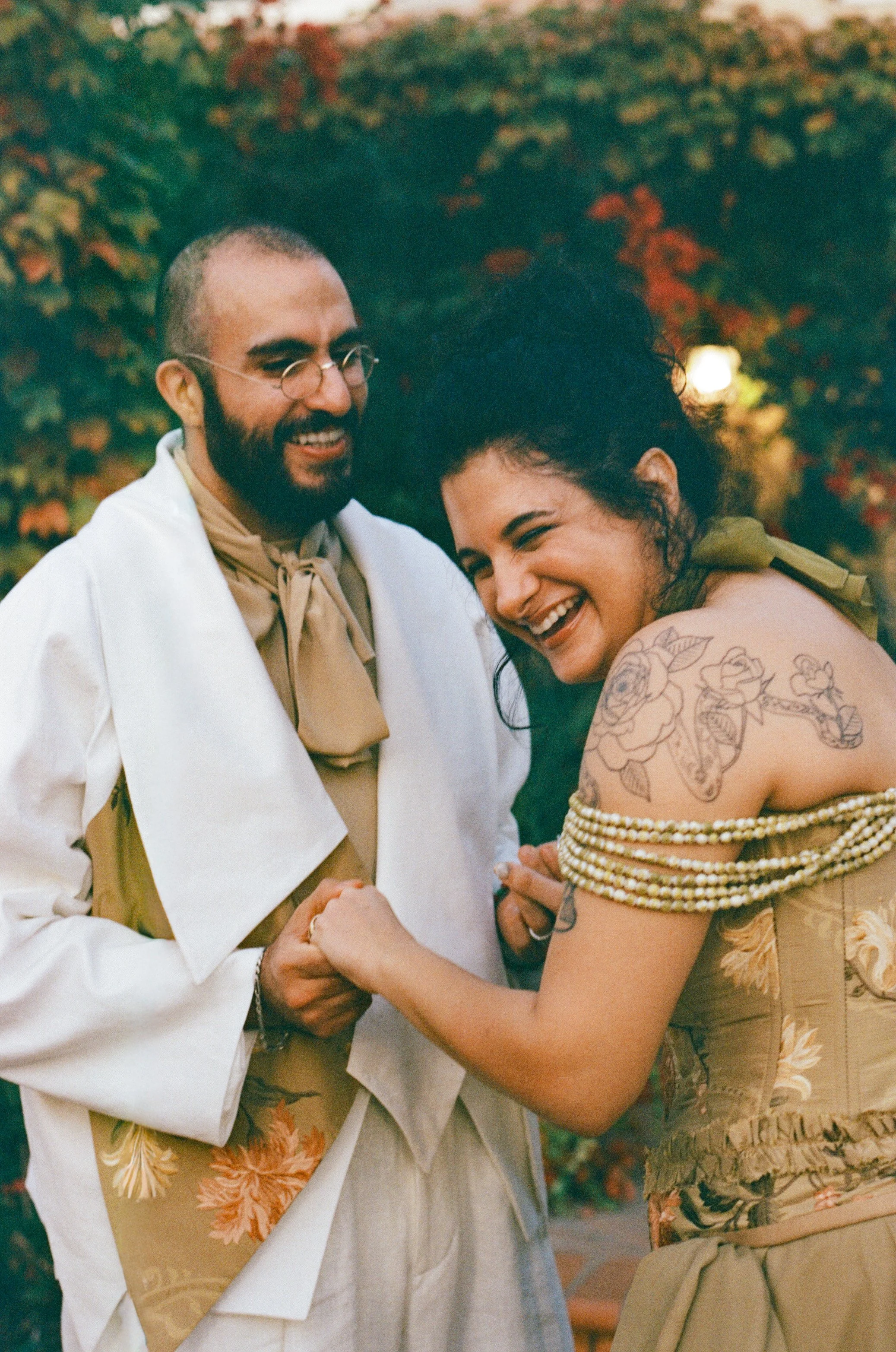 A man with glasses and a beard and a woman with curly hair and tattoos are smiling and holding hands, dressed in floral and light-colored clothing, outdoors with autumn foliage in the background.