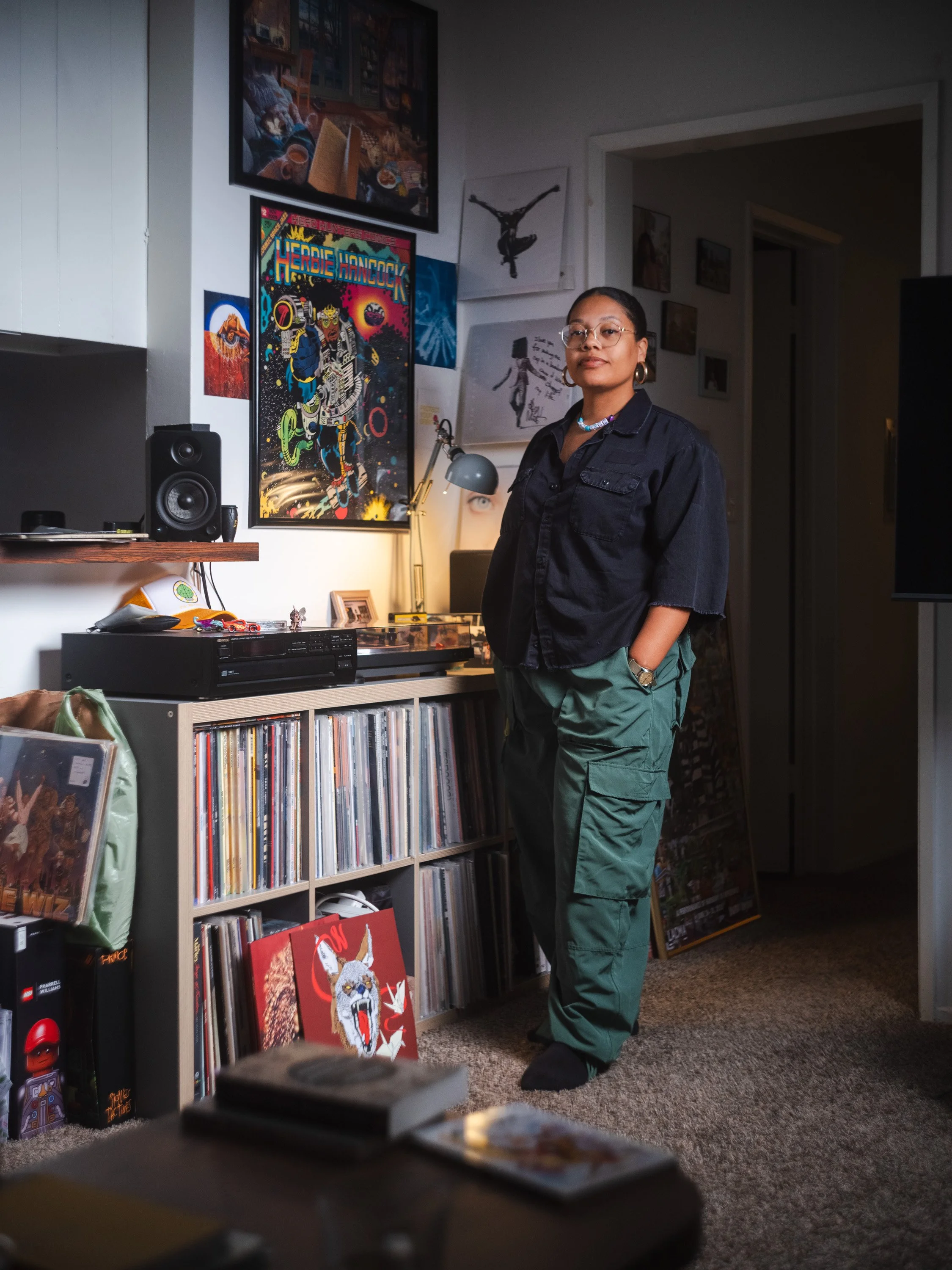 A woman standing in a room decorated with framed artwork, posters, and a collection of vinyl records. She is wearing glasses, a dark shirt, green cargo pants, and hoop earrings. There are speakers, a desk lamp, and various items on the shelves around