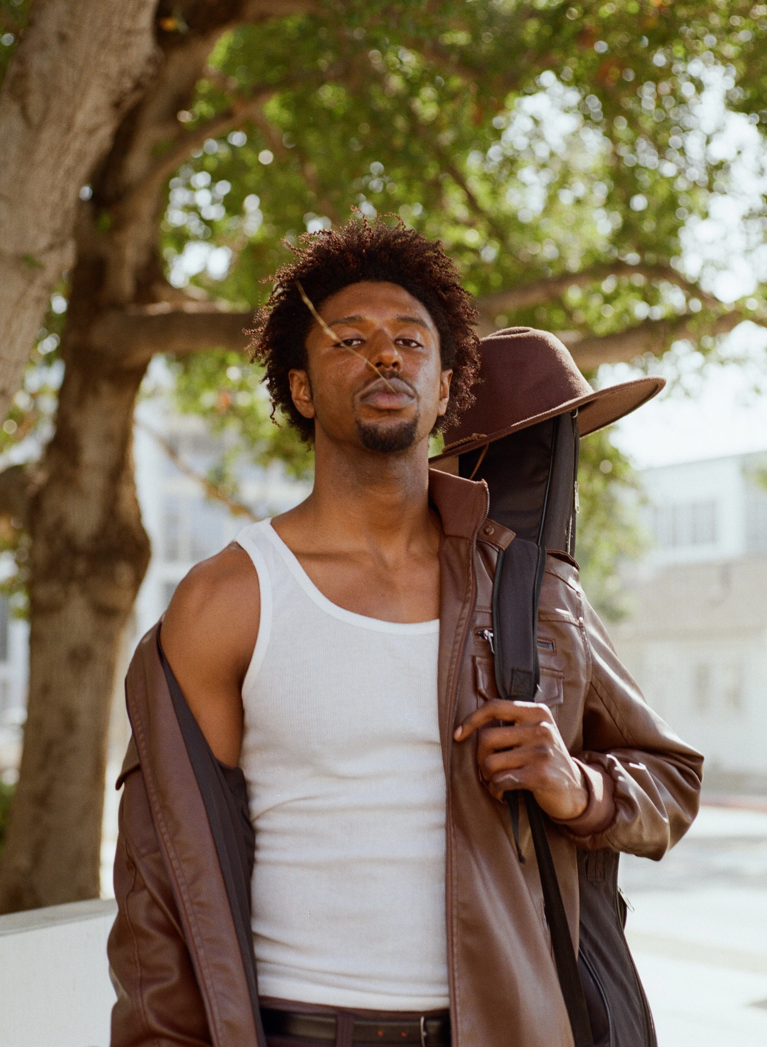 A young man with curly hair, wearing a white tank top and brown leather jacket, standing outdoors near a tree, holding a backpack strap over his shoulder, with buildings in the background.