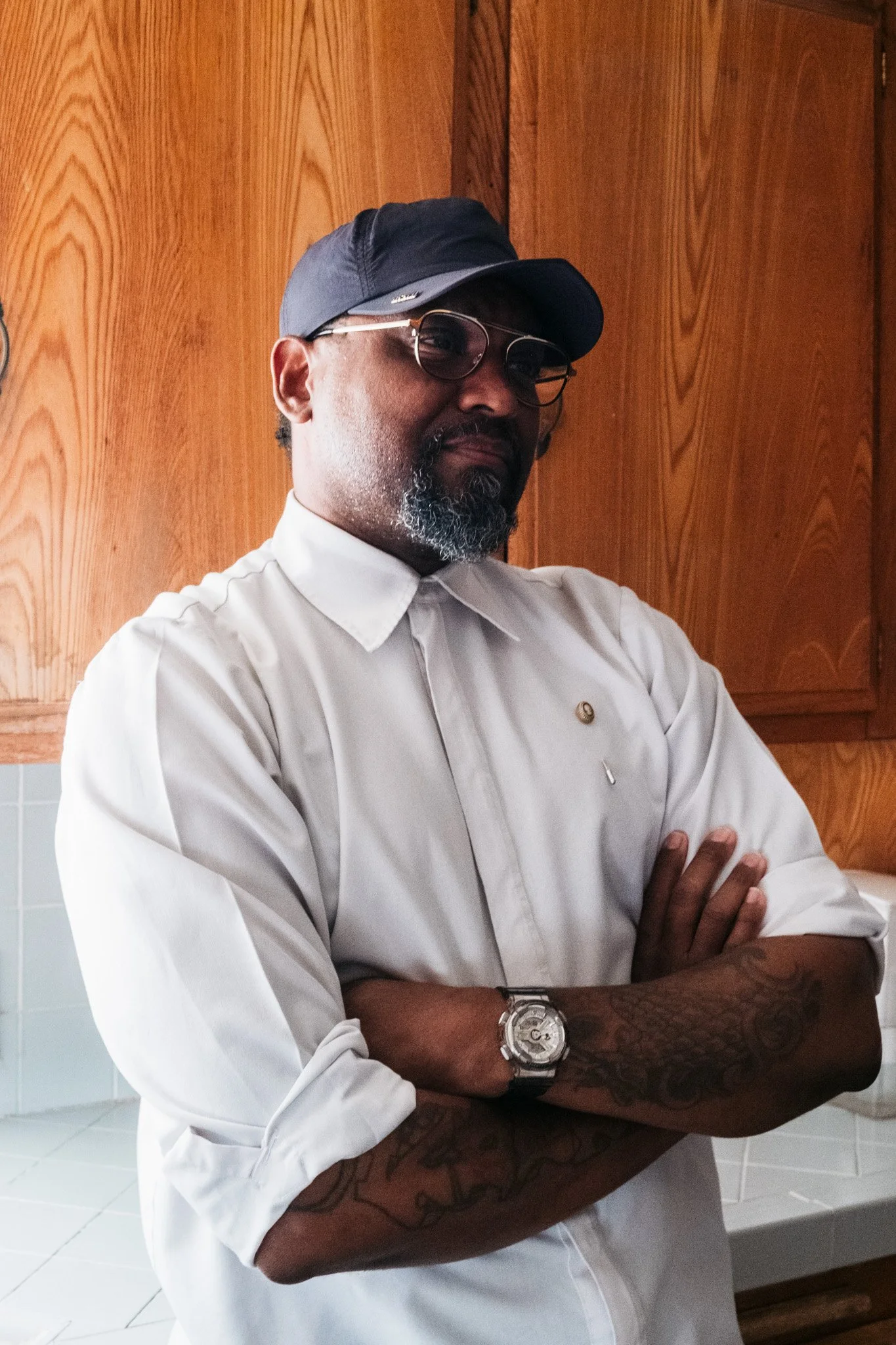 A man with a beard wearing a gray baseball cap, glasses, a white chef's coat, and a large watch, standing with arms crossed against a wooden kitchen cabinets background.