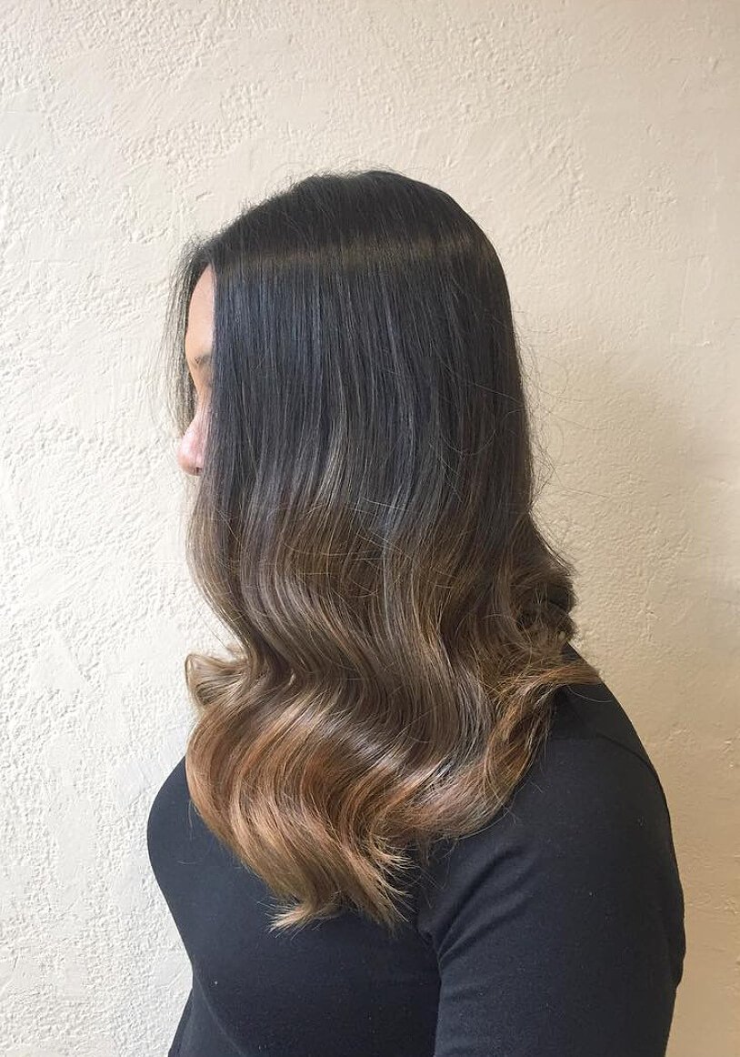 Side view of a woman with long, wavy, dark brown to light brown ombre hair, standing against a textured beige wall.