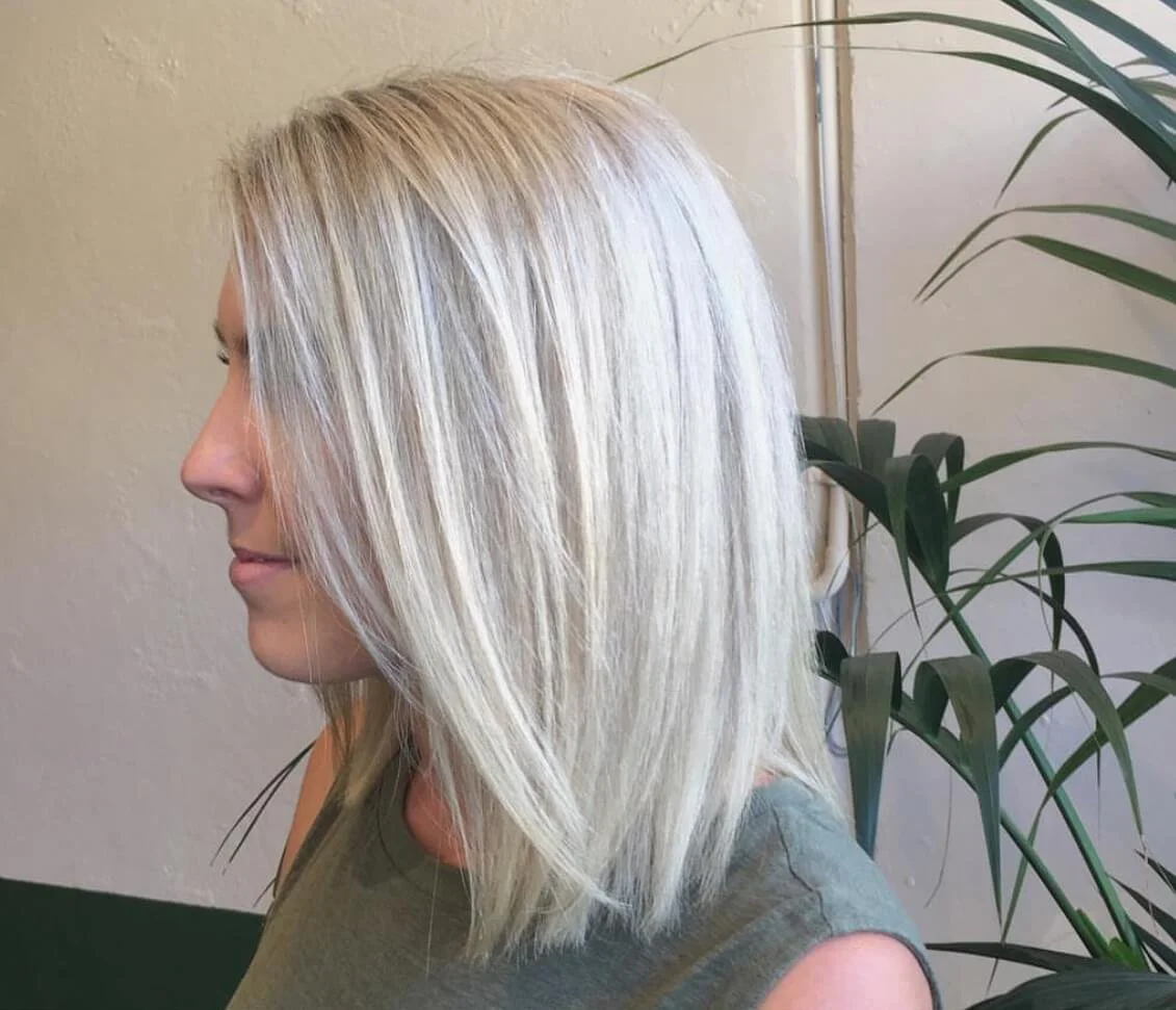 Side view of a woman with shoulder-length blonde hair, standing next to a green plant on a beige wall background.