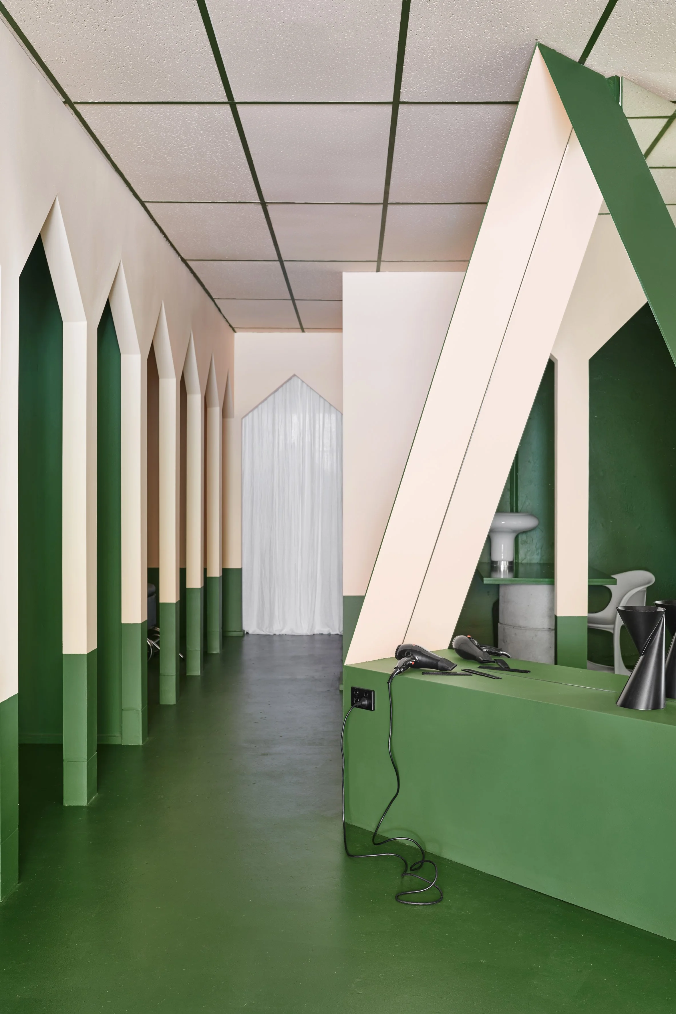 Interior of a modern space with green flooring, green and white geometric wall structures, a white fabric curtain at the end, and a green counter with hair styling tools like a hairdryer and flat iron.