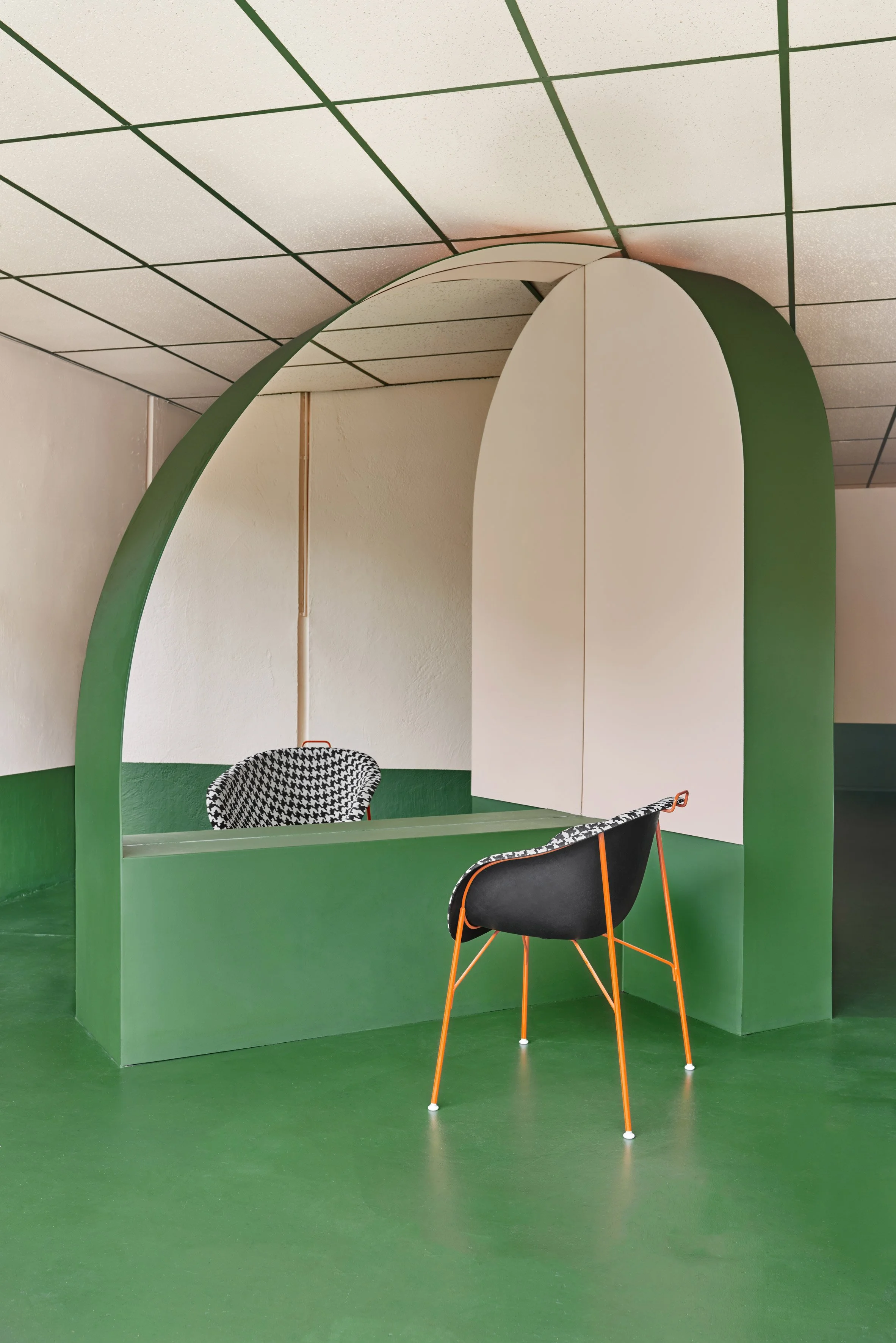 Modern interior with a green curved partition, white walls, a black chair with orange legs, and patterned chairs, under a ceiling with grid-patterned tiles.