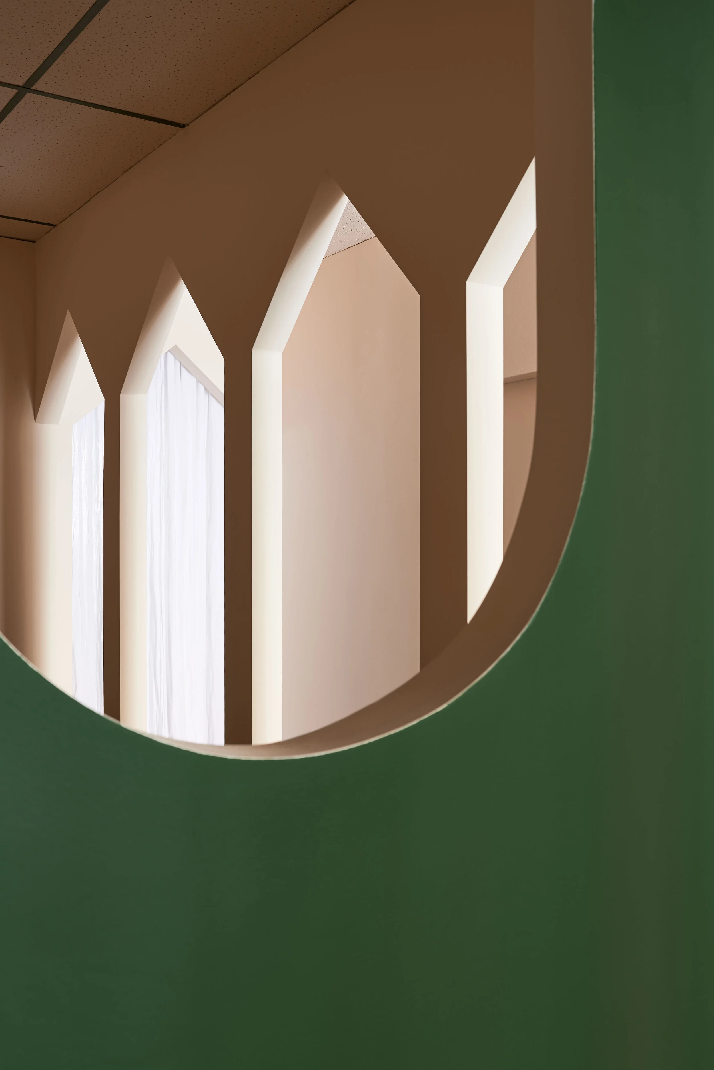 Close-up of interior architectural feature with a cut-out view showing white vertical beams or columns through an oval shaped opening in a green wall, with beige ceiling panels.