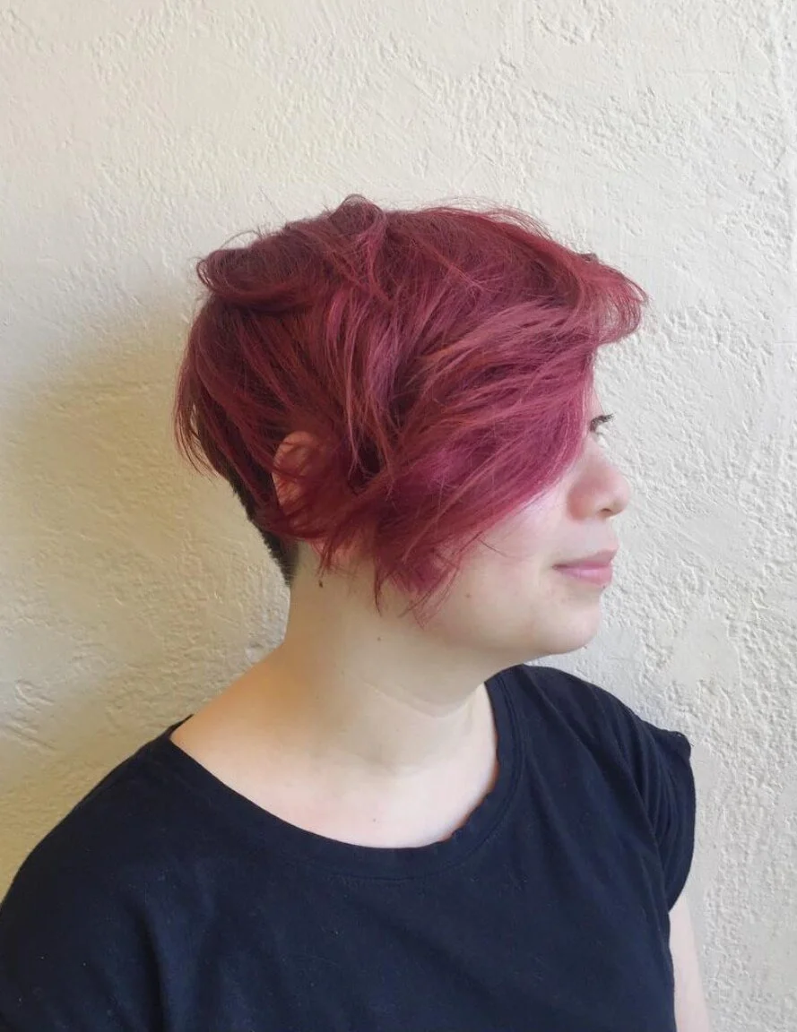 Side profile of a person with short, wavy, burgundy-colored hair wearing a black shirt against a textured beige wall.