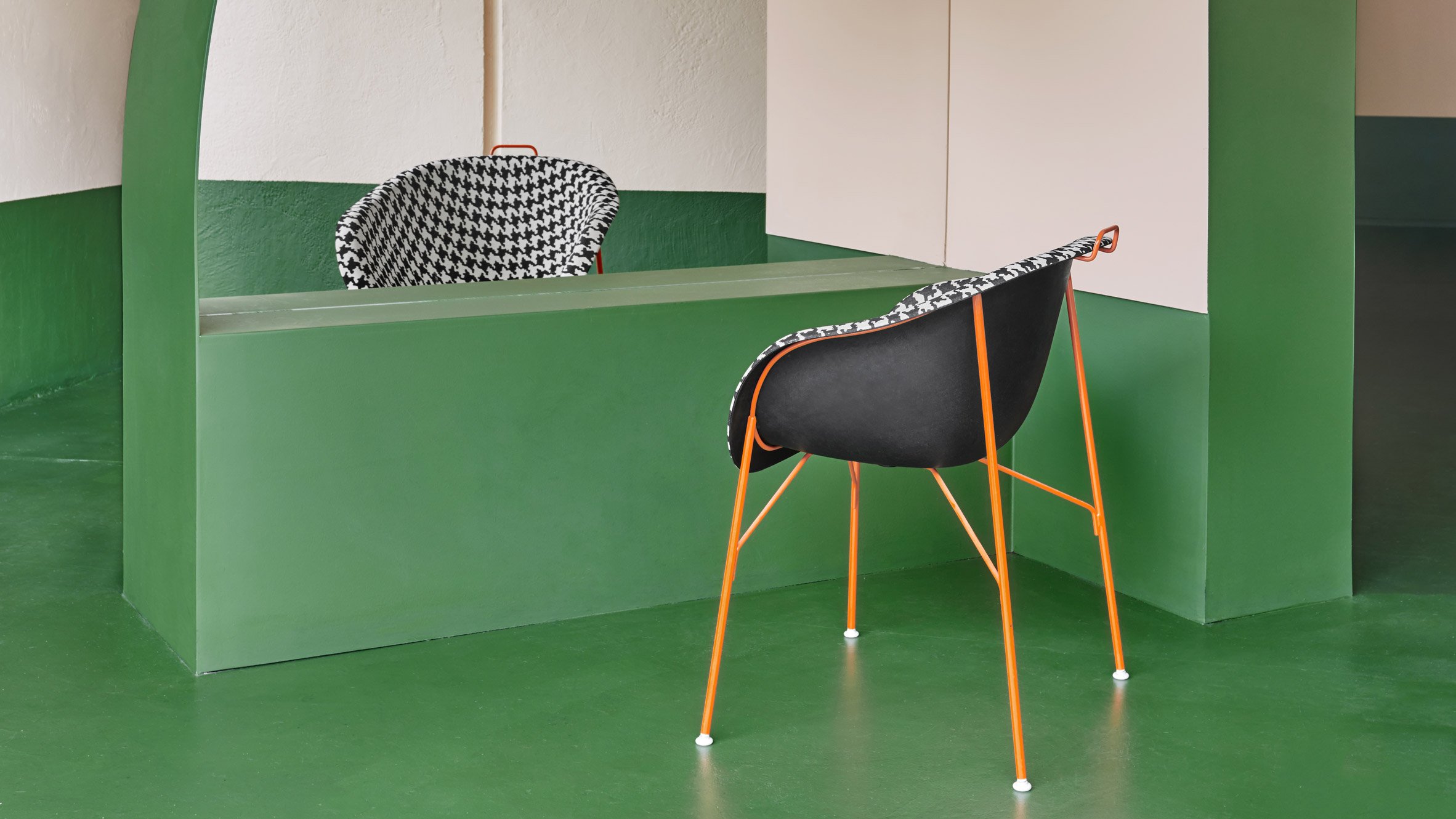 A room with green flooring and walls, featuring a green counter with a mirror and two chairs with black seats and houndstooth-patterned fabric backs. One chair has orange metal legs, and the other has black. There is a white wall with a small cupboar