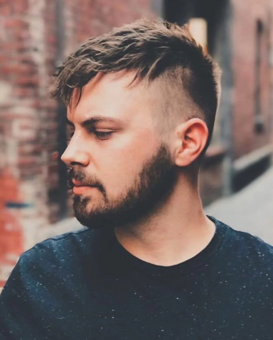 Side profile of a young man with a styled haircut and a beard, standing outdoors with a blurred brick building background.
