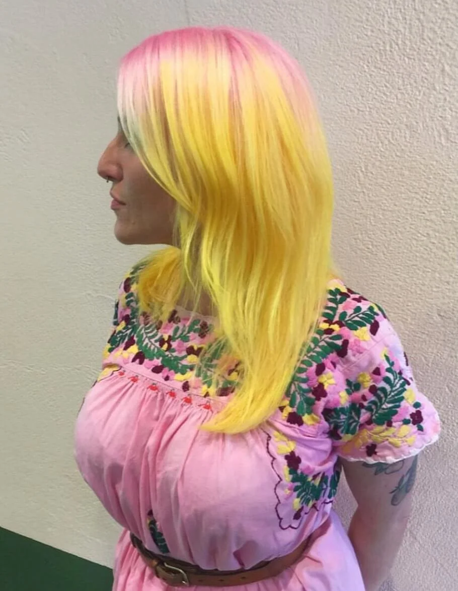 Profile of woman with multi-colored hair in pink, yellow, and blonde shades, wearing a pink embroidered dress with floral patterns.
