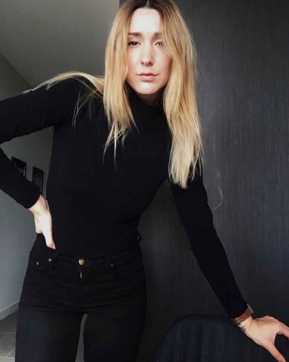 A woman with long blonde hair wearing a black turtleneck and black pants, leaning on a dark textured wall with one hand on her hip and the other resting on furniture.