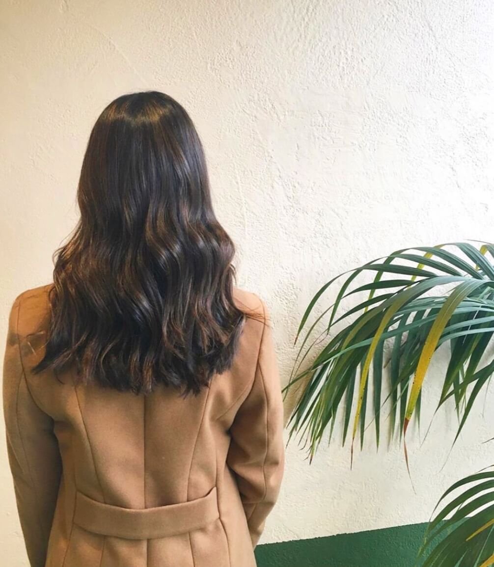 A woman with wavy brown hair wearing a brown coat, standing near a potted plant against a textured beige wall.