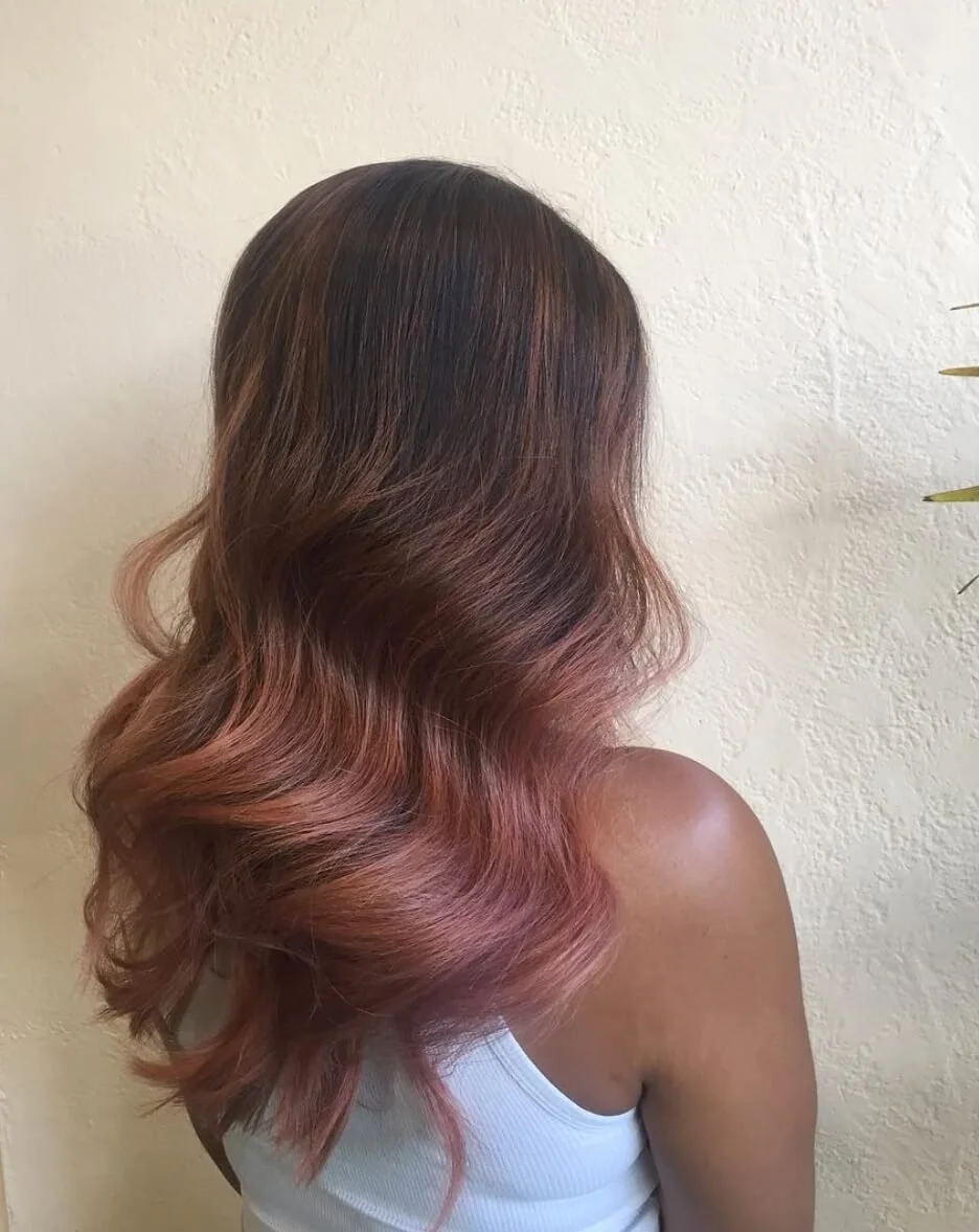 Side view of a woman with wavy, ombre hair transitioning from dark brown at the roots to pink at the ends, wearing a white sleeveless top against a cream-colored textured wall.