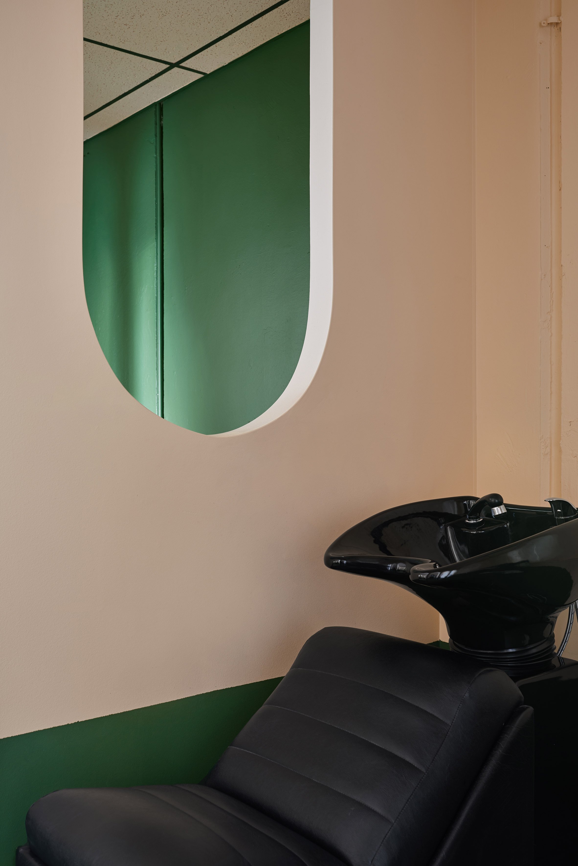 Hair salon or barber shop with a black shampoo sink and a black reclining salon chair. The wall behind has a large round mirror with a white frame, and the wall is painted beige with dark green baseboard and ceiling trim. The floor is green.