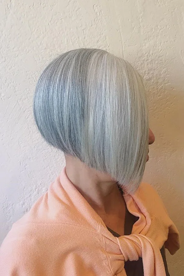 A woman with a stylish, asymmetrical short bob haircut featuring silver-gray hair color, wearing a peach-colored top against a textured beige wall.