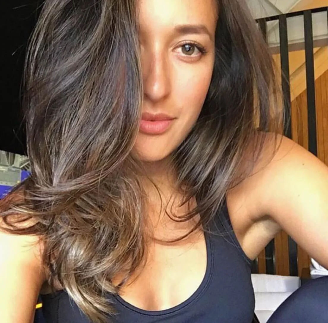 A woman with long, wavy brown hair partially covering her face, wearing a black sleeveless top, with a wooden background.