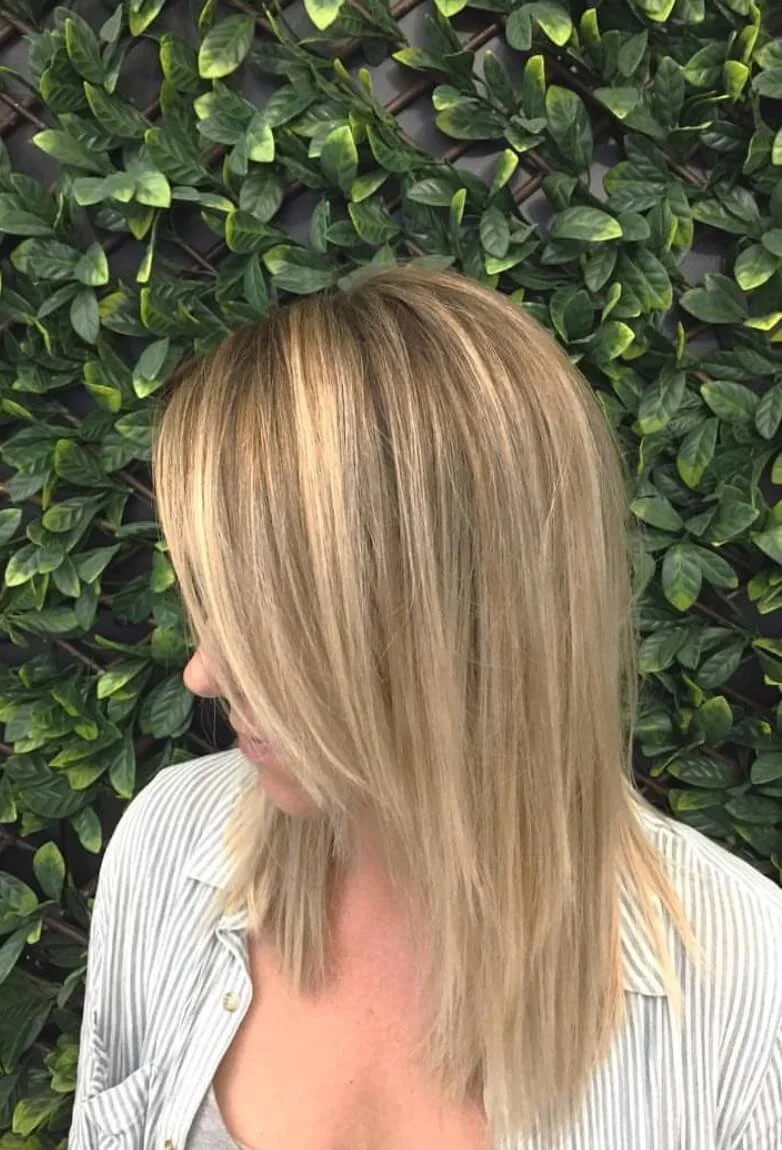 A woman with blonde hair, wearing a striped shirt, standing in front of a green leafy background.