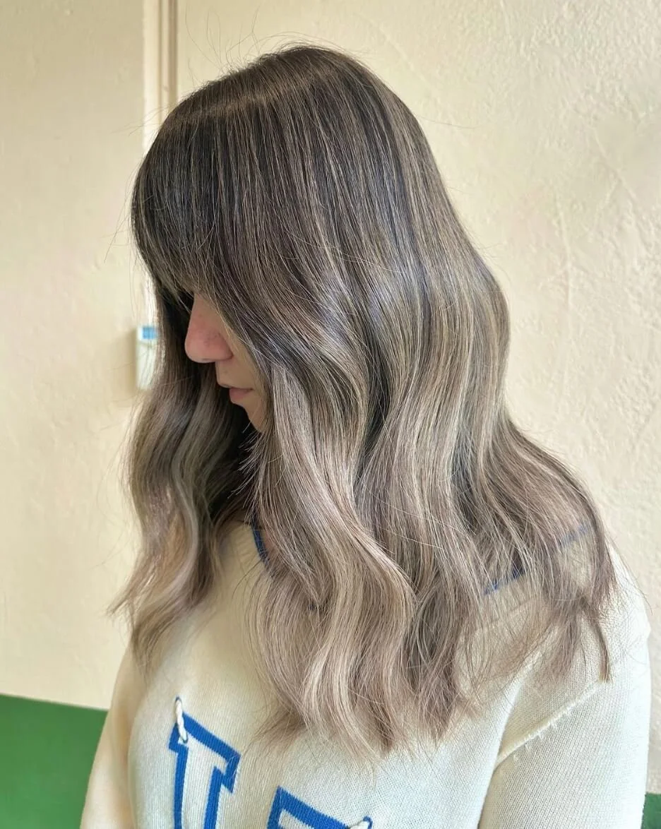 Side view of a woman with wavy, ombre hair transitioning from dark brown at the roots to light blonde at the ends, standing against a beige wall.