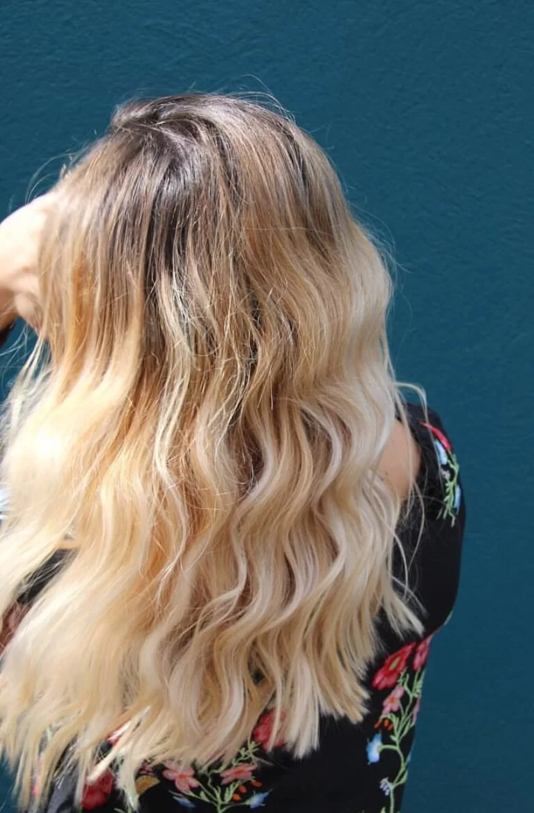 Woman with long, wavy blonde hair, wearing a black floral top, standing against a blue wall.