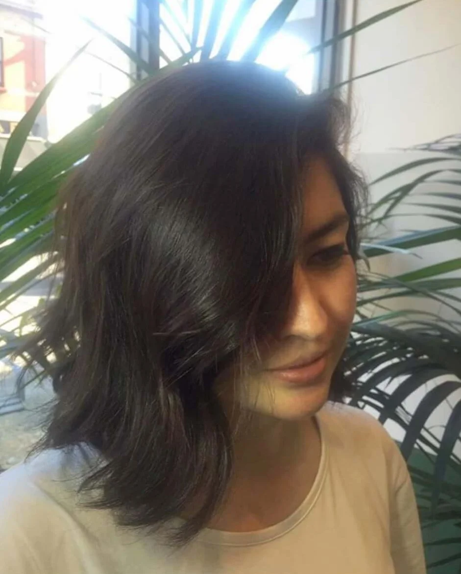 A woman with shoulder-length, wavy, dark hair standing near green indoor plants, with sunlight coming through a window behind her.