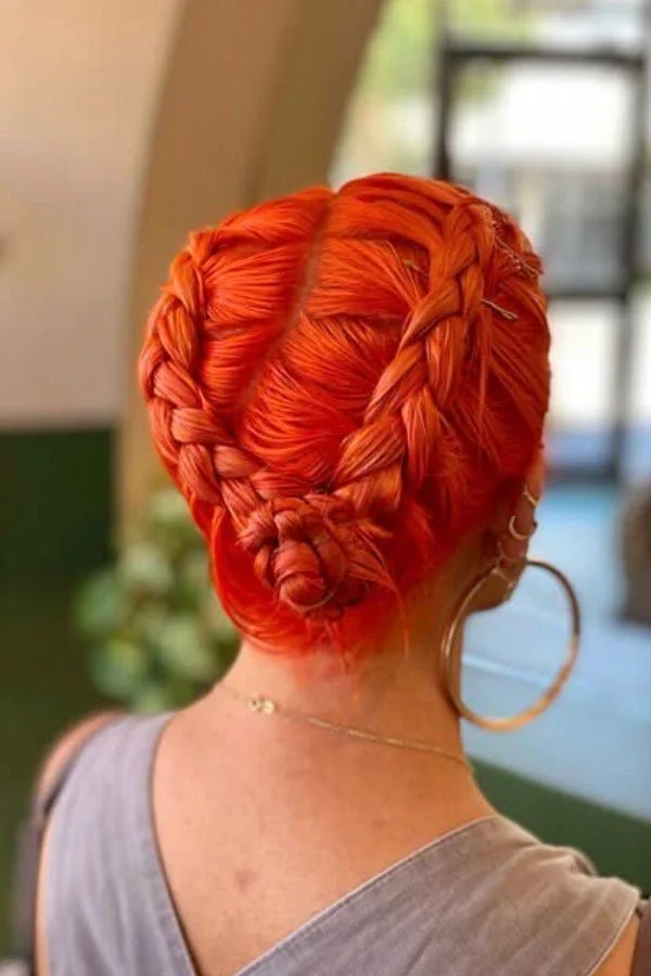 Back of a woman with bright orange hair styled in elaborate braids and buns, wearing large hoop earrings and a tank top.