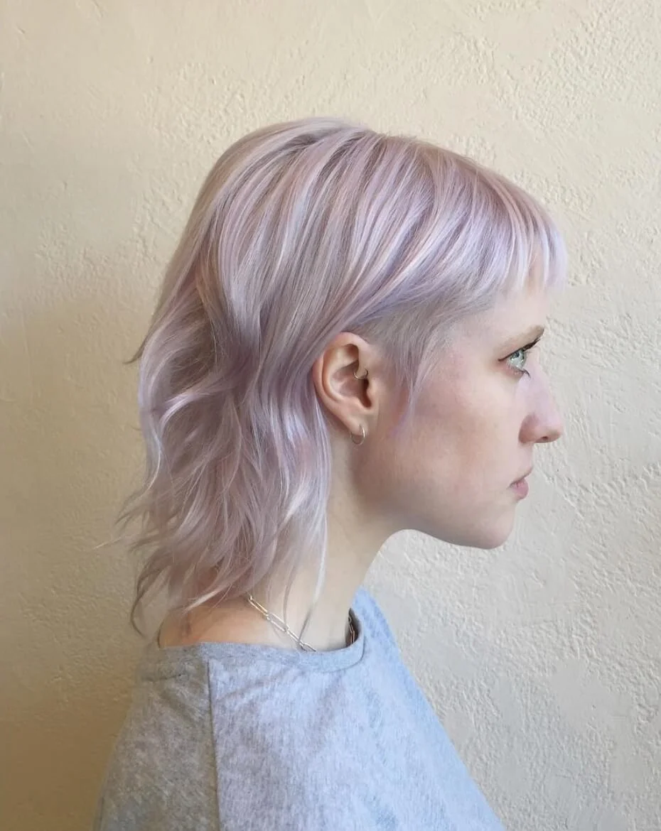 Side profile of a woman with short, wavy, light lavender hair against a beige wall.