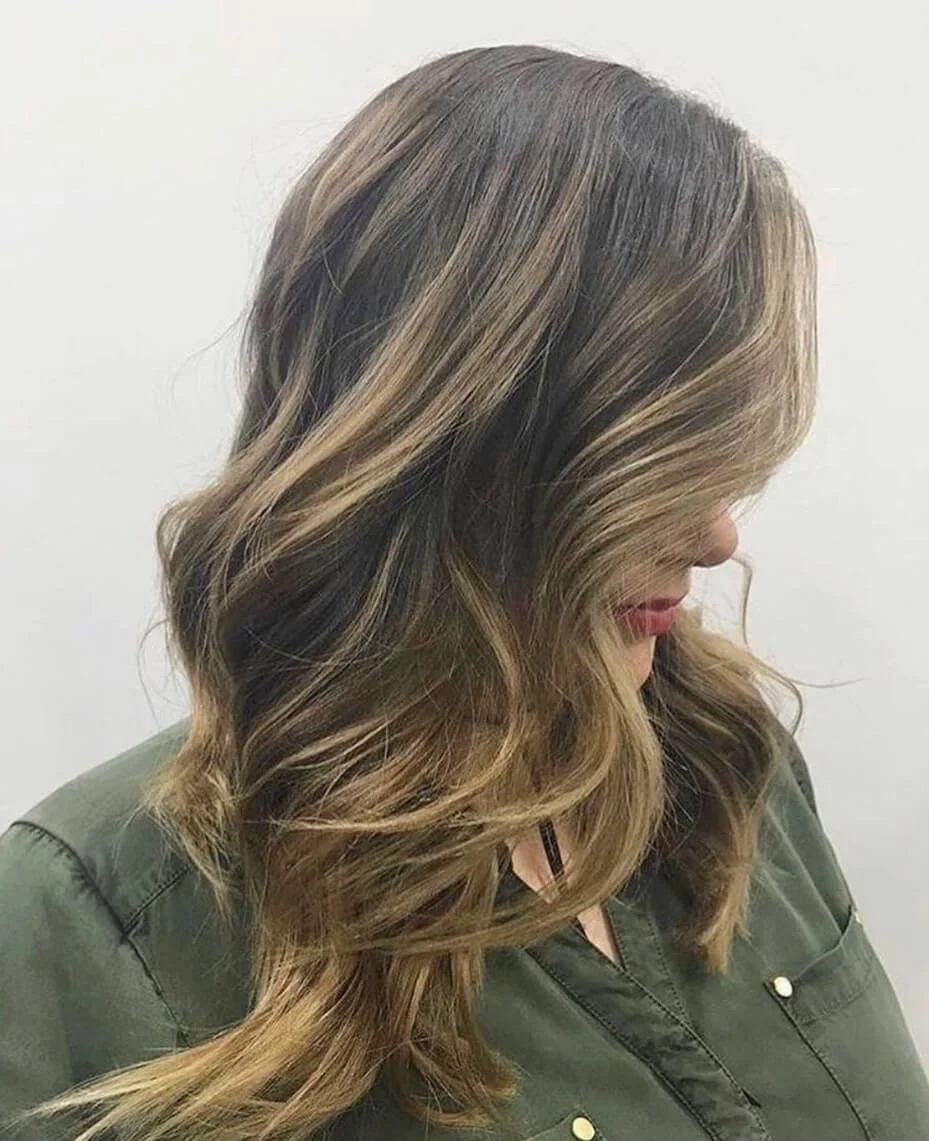 Woman with wavy, shoulder-length hair with highlights, wearing an olive green jacket, looking downward against a plain white wall.