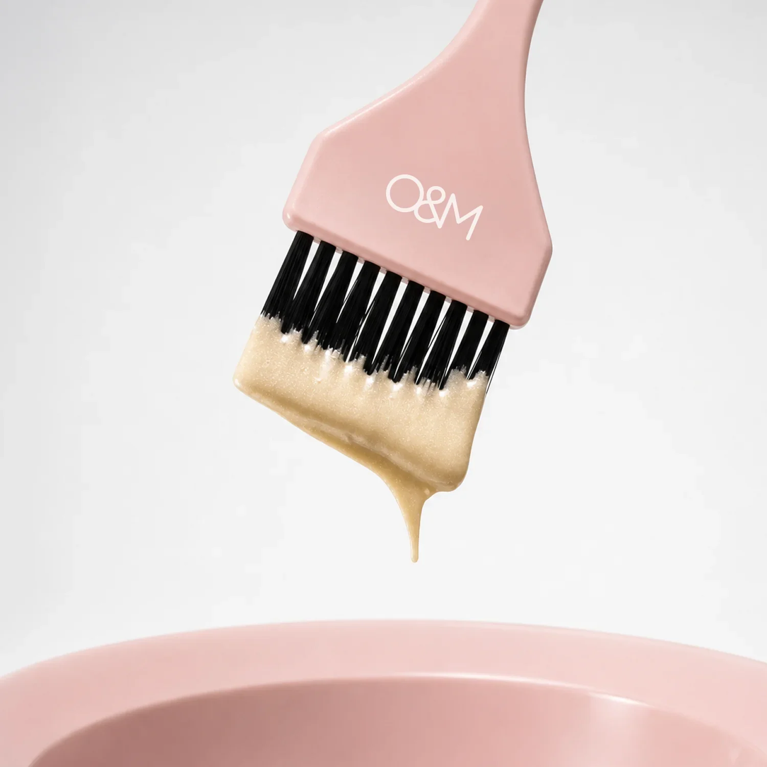 A close-up of a pink and beige mini cleaning brush with black bristles, cleaning the edge of a pink container, with a beige liquid dripping from the brush.