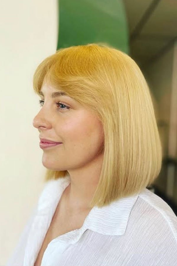 Side profile of a woman with shoulder-length blonde hair, wearing a white shirt, standing indoors.