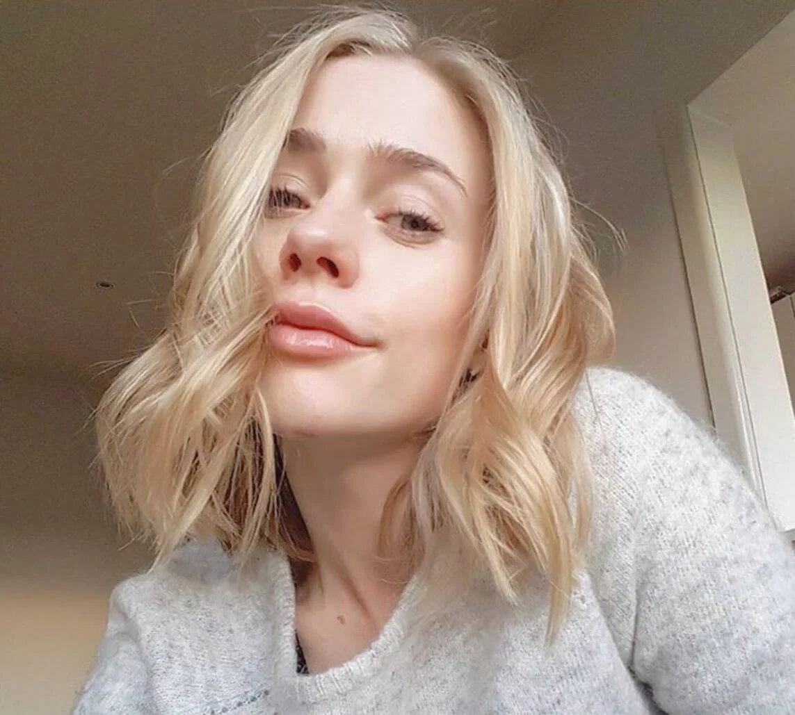 Close-up of a woman with blonde wavy hair, wearing a light-colored top, with a neutral indoor background.