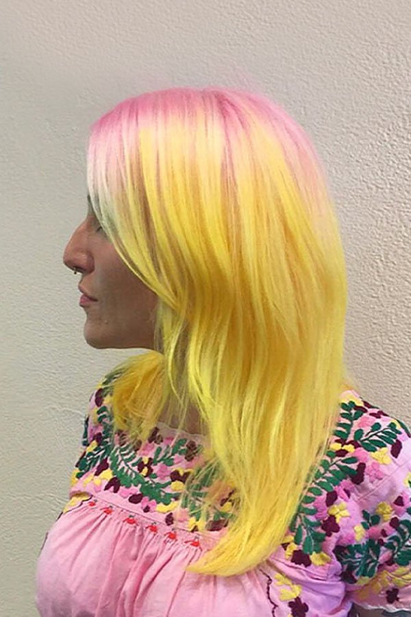 Side view of a woman with shoulder-length hair dyed in pastel pink, yellow, and green, wearing a pink embroidered top with floral patterns.