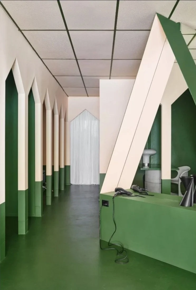 Interior of a building with a green and beige color scheme, featuring arched wall details, a white curtain at the end of a hallway, a sink, and hair styling tools on a counter.
